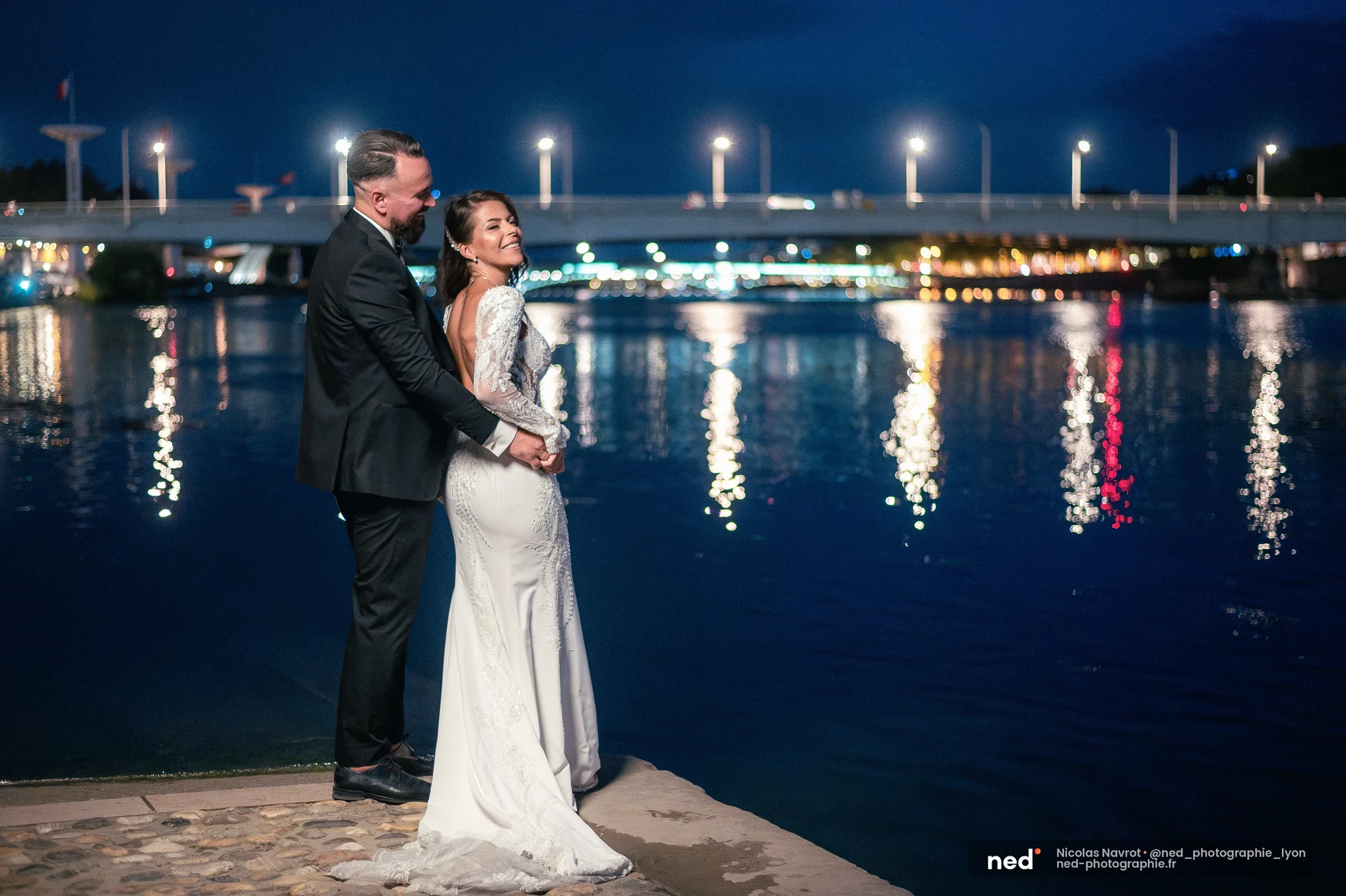 Couple de mariés enlacés au bord du Rhône de nuit, pont illuminé en fond, séance photo couple à Pl. de l'Hôpital