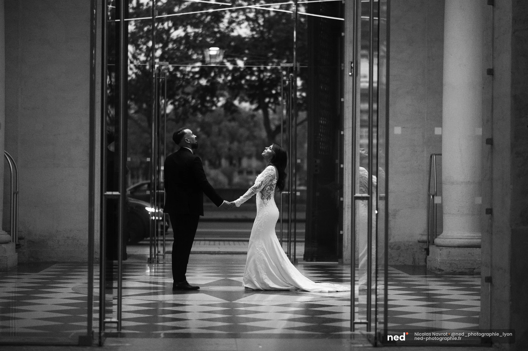Mariés se tenant par les mains dans un hall en verre, sol en damier, noir et blanc, séance photo couple à Pl. de l'Hôpit