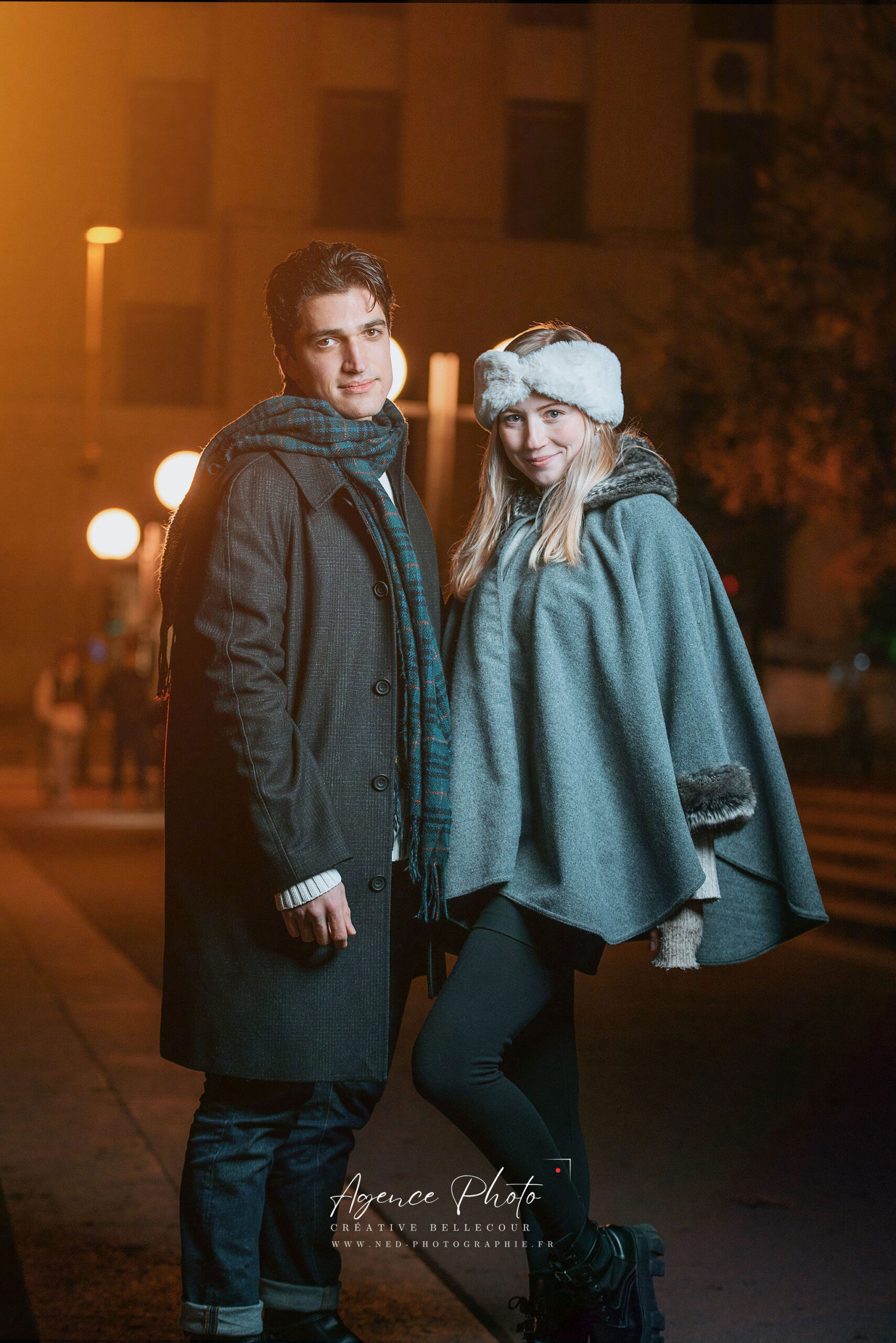 Jeune couple sourit à la caméra dans une rue pavée la nuit, éclairés par les réverbères dorés d'une façade Renaissance