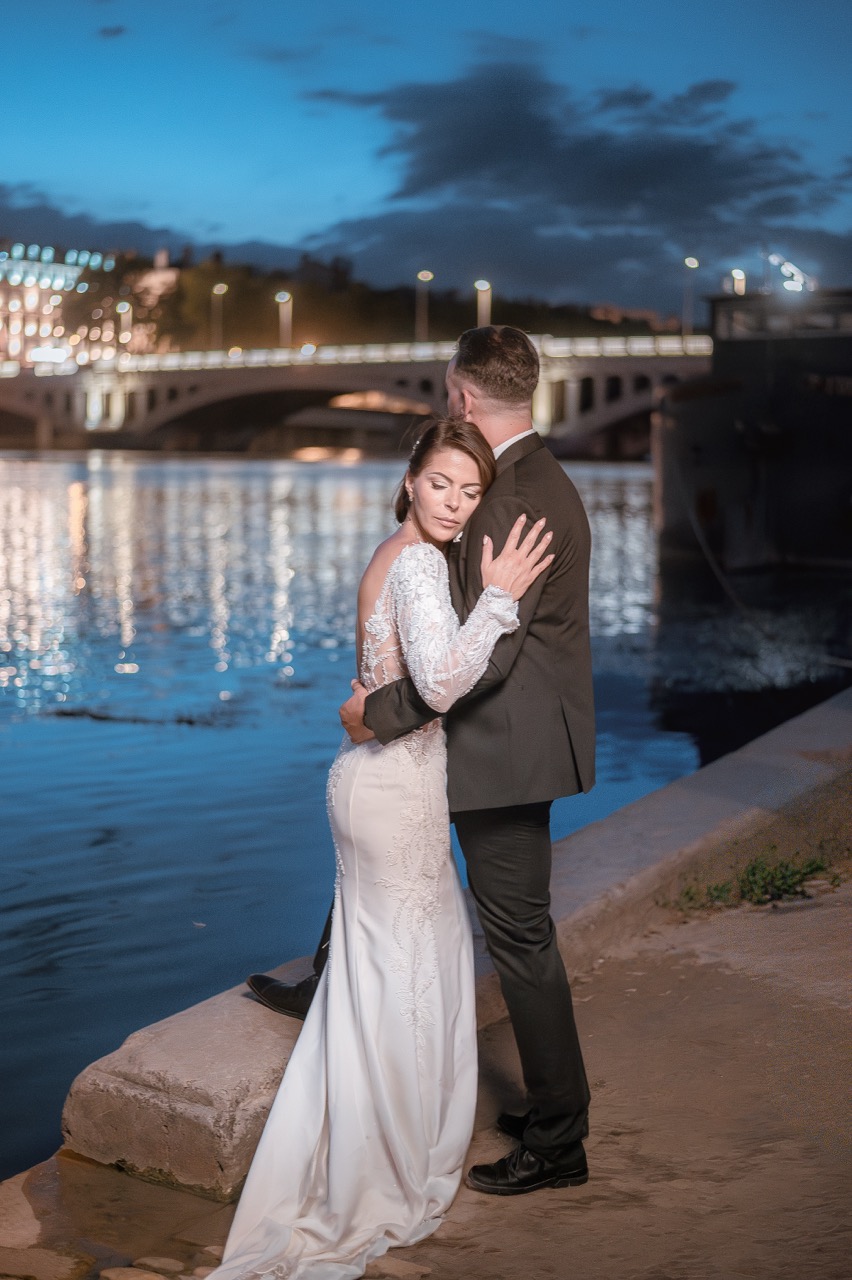 Mariée en robe blanche brodée et marié en costume sombre enlacés devant le pont illuminé de Lyon au crépuscule