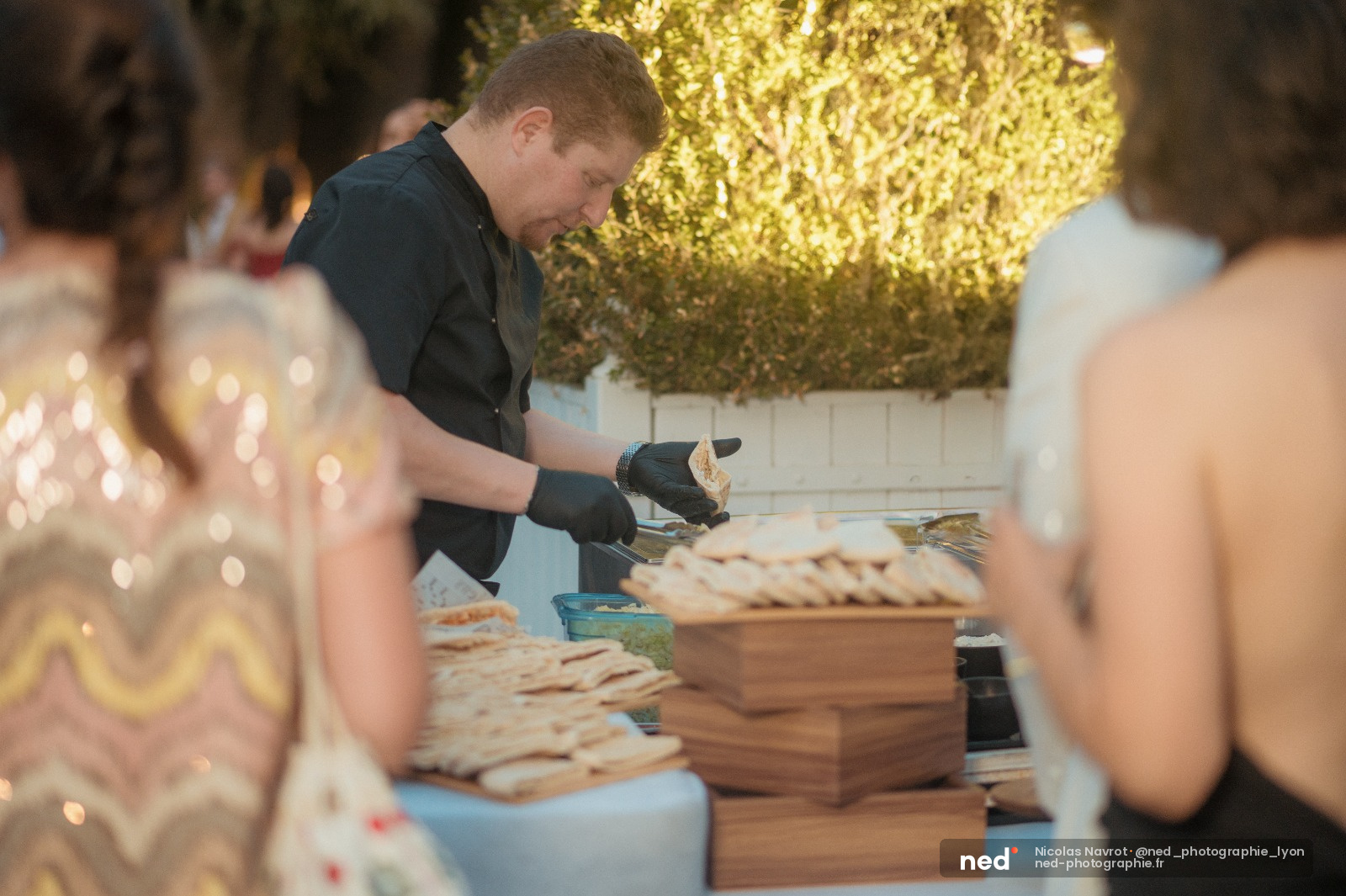 Jeune serveur en costume noir et gants prepare les assiettes de pain lors d'un banquet en exterieur, sous une decoration lumineuse