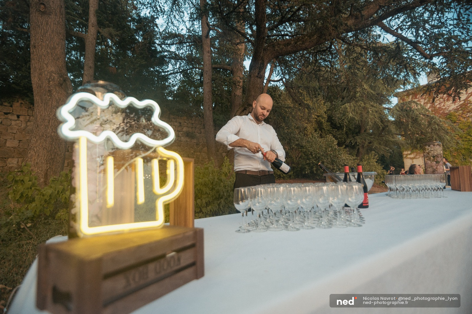 Homme en chemise blanche versant du vin à une table de réception en extérieur, enseigne néon bière en premier plan