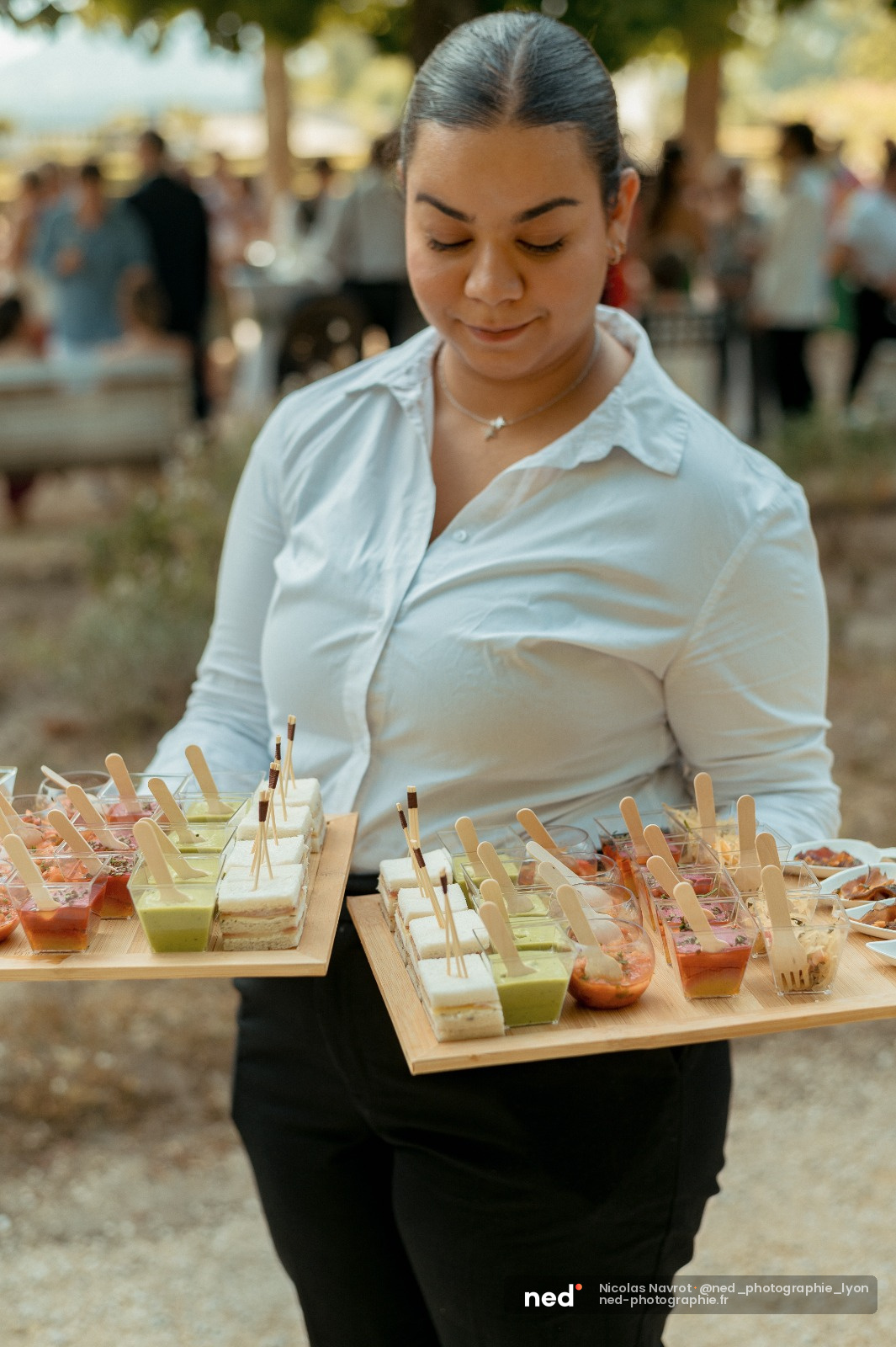 Serveuse en chemise blanche présente des petits fours et verrines colorées sur plateaux en bois lors d'un événement en extérieur
