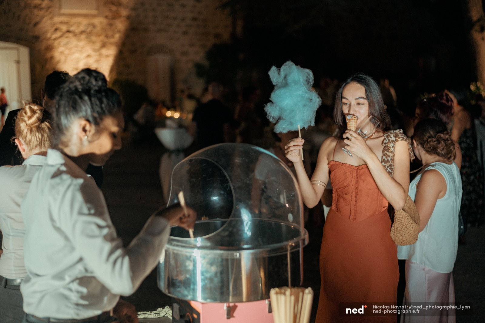 Jeune femme en robe terracotta tenant une barbe à papa turquoise lors d'une fête en extérieur la nuit, entourée d'autres invités