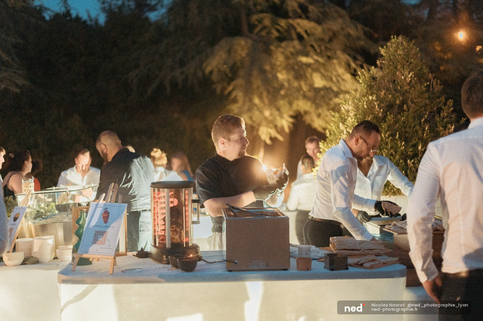 Groupe de personnes autour d'une table lors d'une soirée en extérieur la nuit, éclairage chaud et végétation en arrière-plan