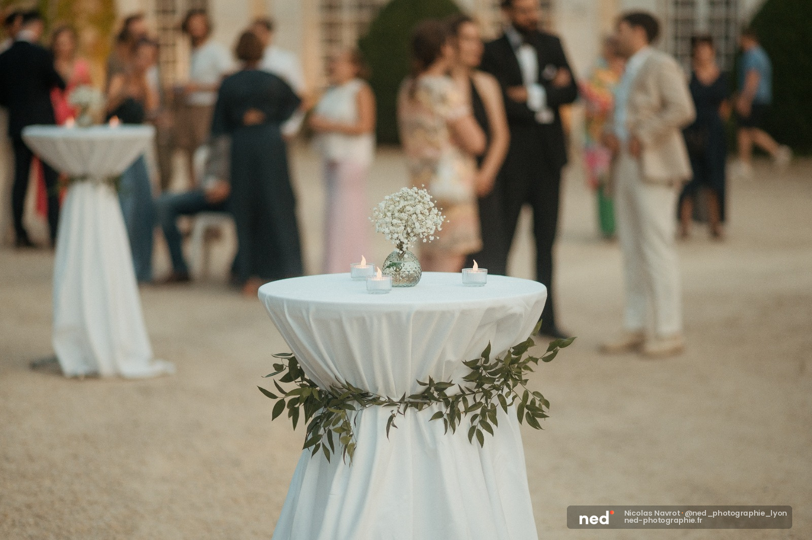 Table ronde avec nappe blanche, vase de gypsophile et bougies, invités en arrière-plan lors d'une réception de mariage