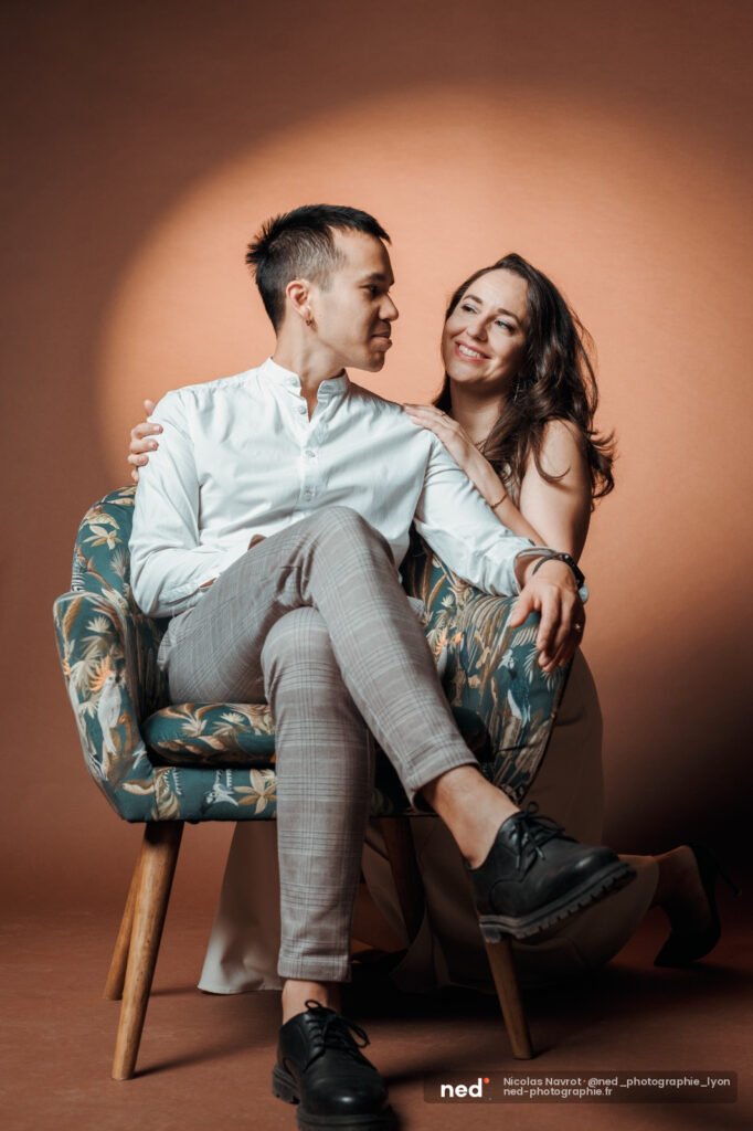 Jeune couple assis sur un fauteuil, la femme souriante appuyée contre l'homme, fond neutre ocre en studio
