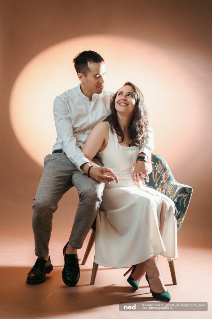 Homme en chemise blanche et femme en robe crème posent assis sur une chaise, souriants, fond neutre tamisé