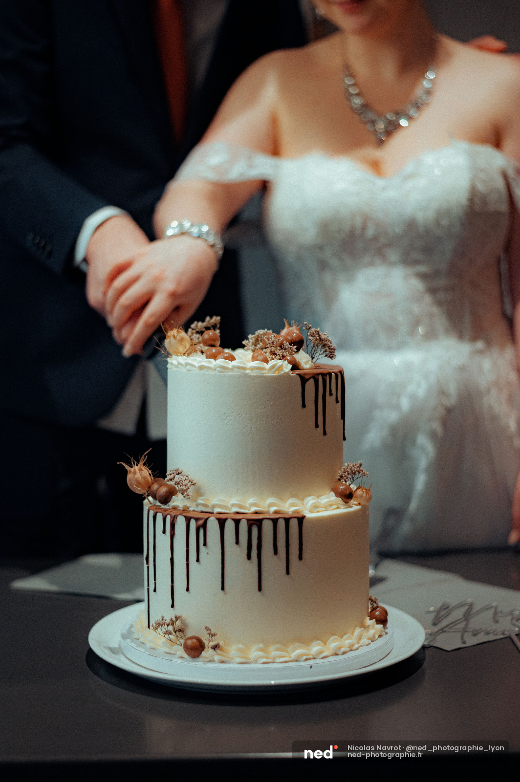 Mains des mariés s'apprêtant à couper un gâteau de mariage à deux étages, glaçage blanc, coulures chocolat et physalis séchés.