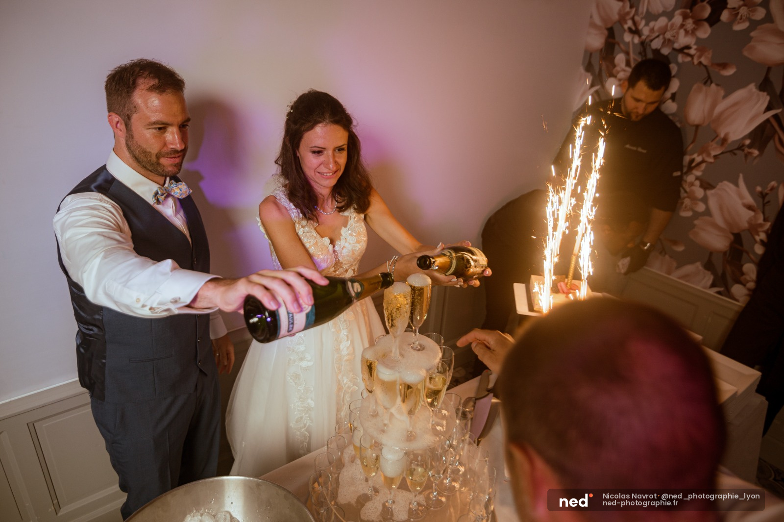 Marié en costume et mariée en robe blanche débouchent une bouteille de champagne devant des invités, fontaine lumineuse en arrière-plan