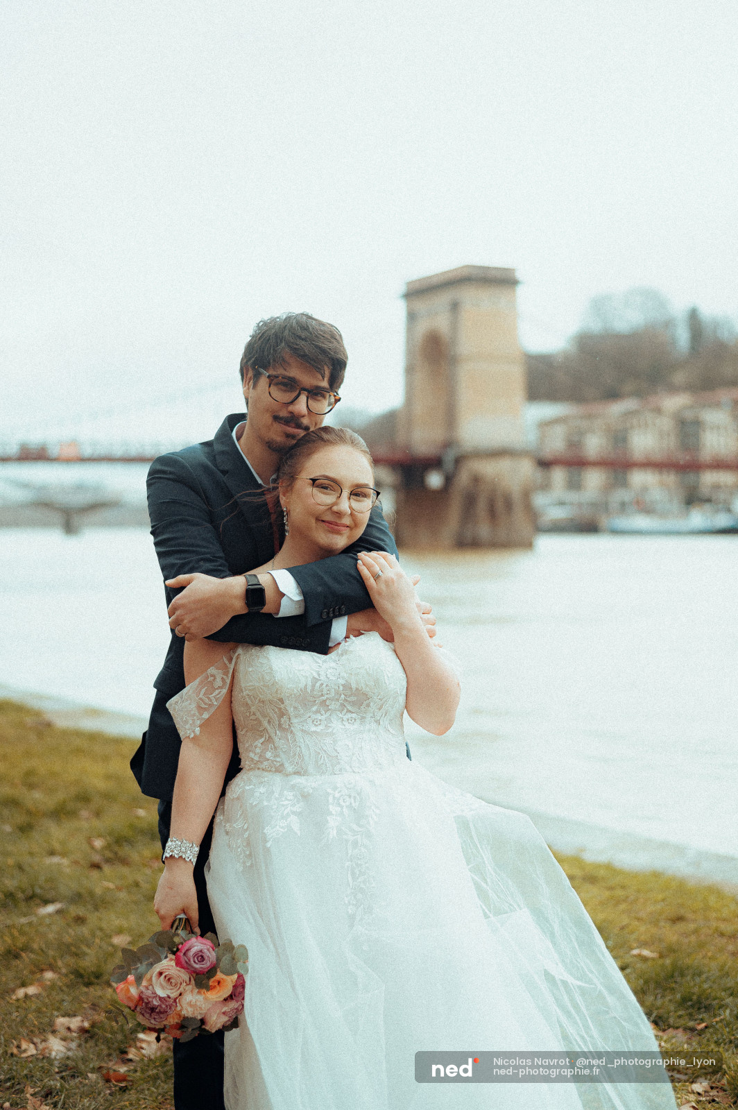 Le marié enlace la mariée par les épaules au bord d'un fleuve, bouquet de roses dans sa main, pont suspendu en arrière-plan.