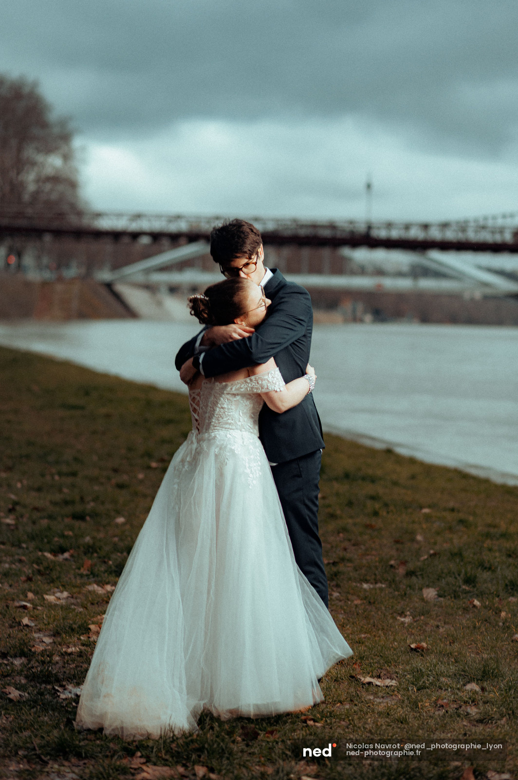 Mariée en robe blanche et marié en costume sombre s'enlacent au bord d'un fleuve, pont métallique visible en arrière-plan, ciel nuageux.