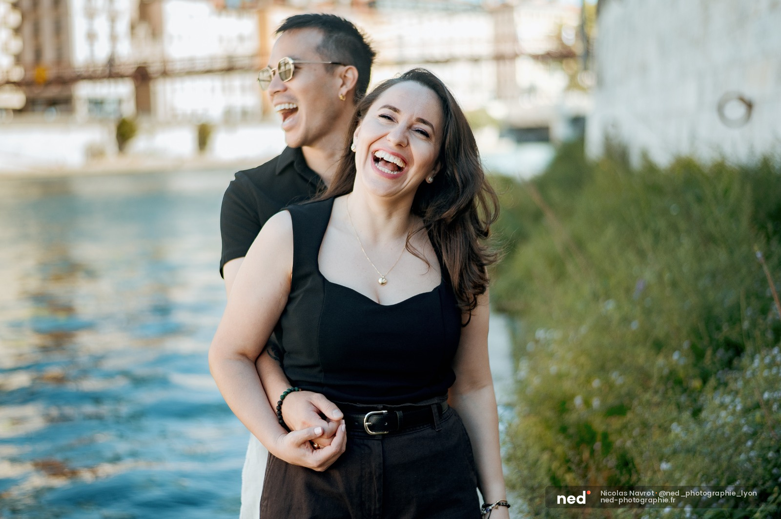 Homme en lunettes et chemise noire enlace une femme en robe noire qui rit, debout près d'une rivière avec des bâtiments en arrière-plan