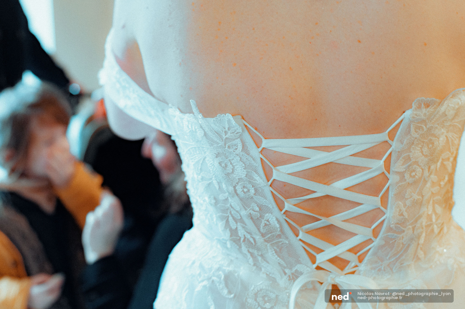 Gros plan sur le dos lacé et brodé d'une robe de mariée en dentelle blanche, avec une silhouette floue en arrière-plan.