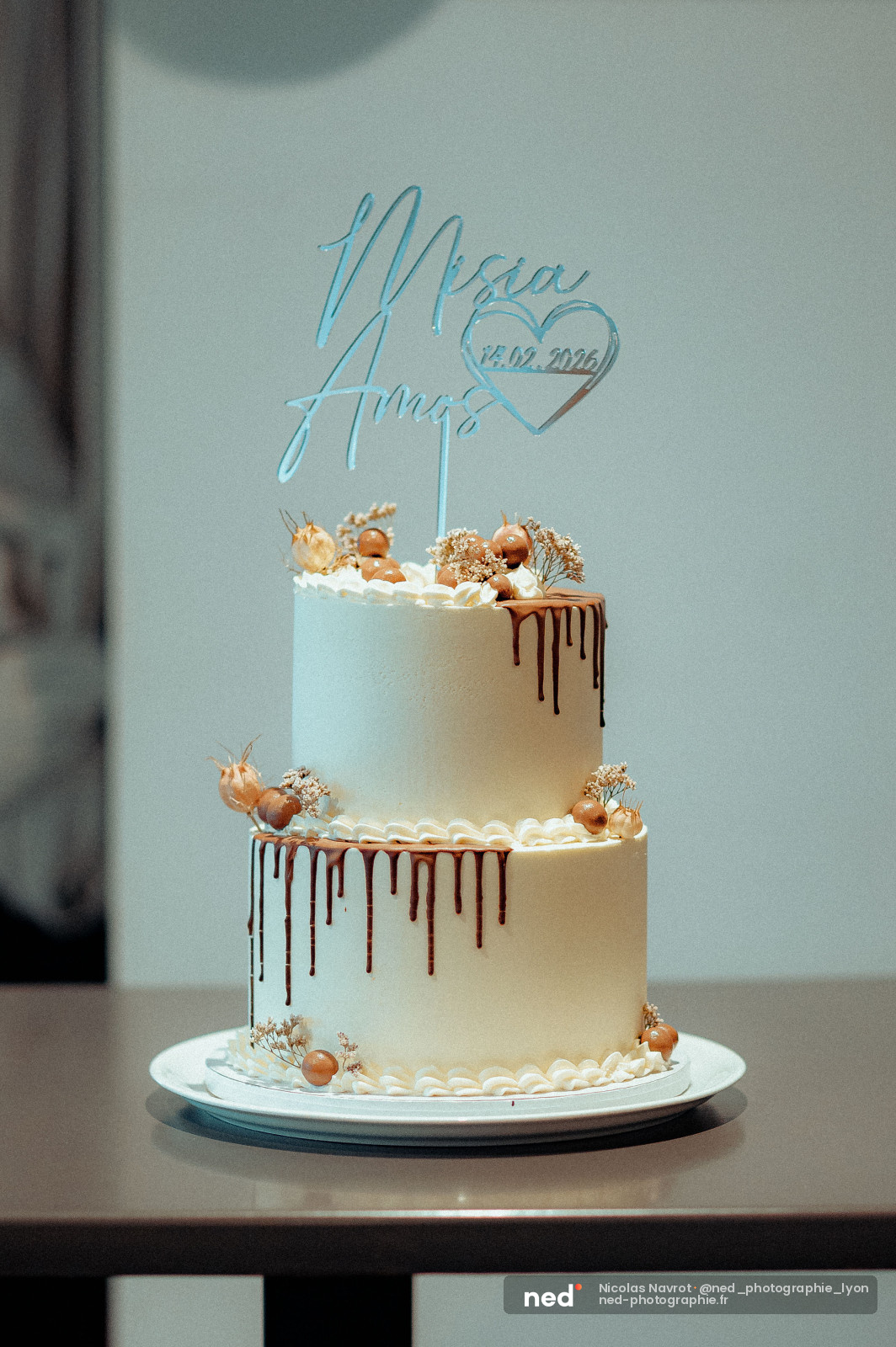 Gâteau de mariage à deux étages, glaçage blanc avec coulures de chocolat, décoré de fleurs séchées et d'un topper acrylique bleu gravé.