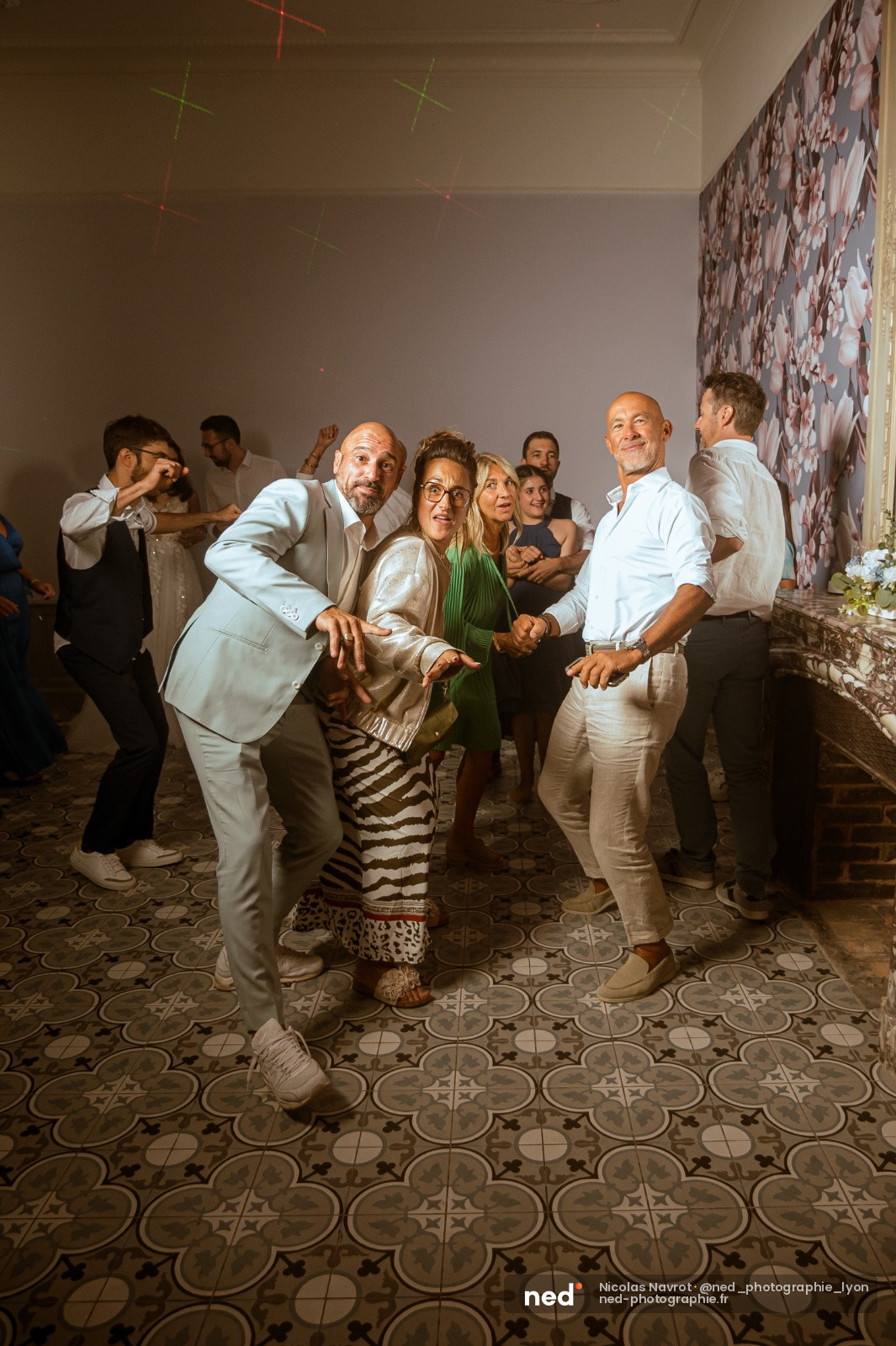 Groupe d'invites dansant et riant ensemble dans une salle de reception, homme en costume clair au centre, carrelage geometrique au sol