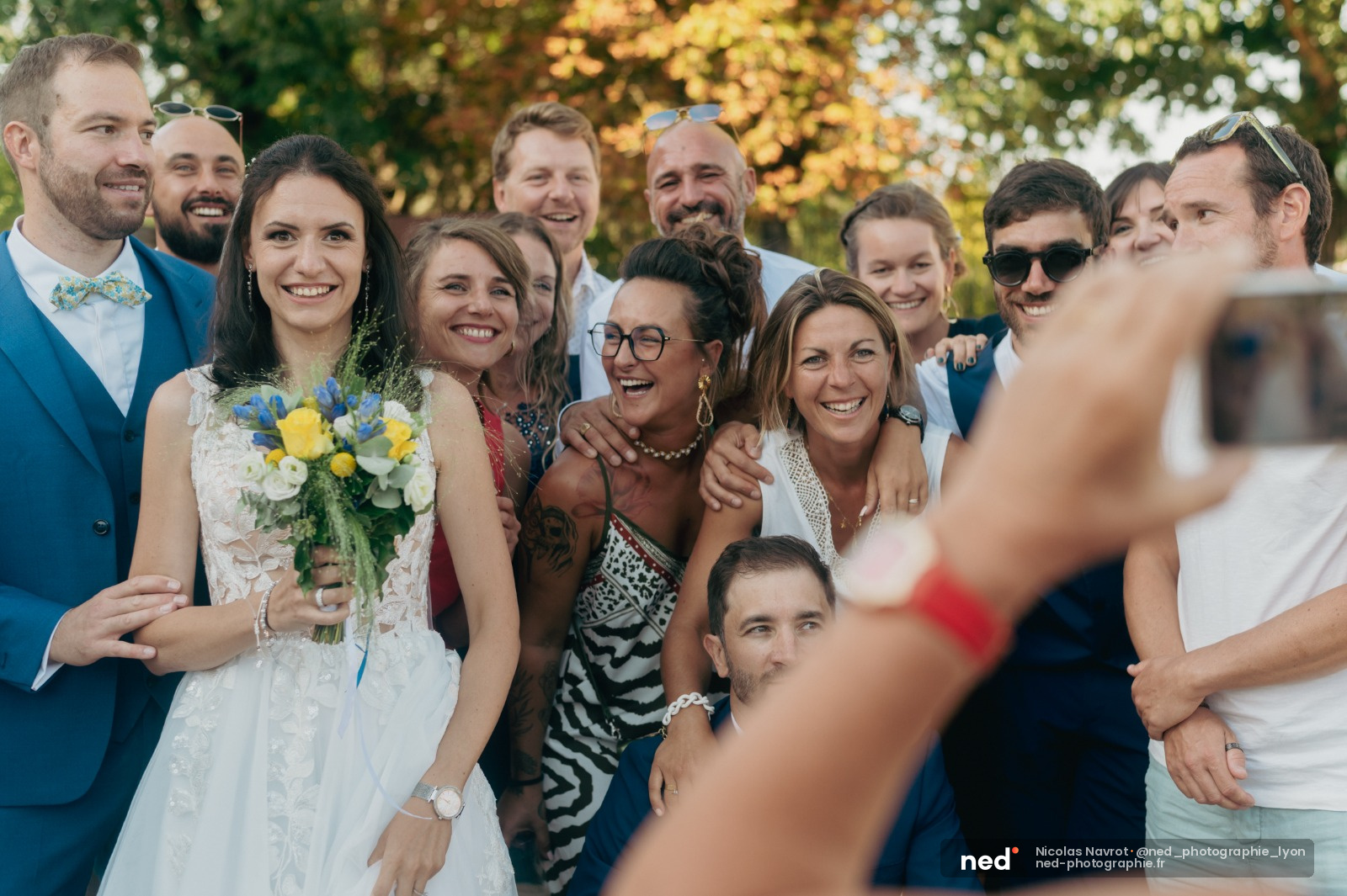 Mariée en robe blanche avec bouquet jaune sourit au milieu d'amis et famille en tenues habillées, feuillage automnal en arrière-plan