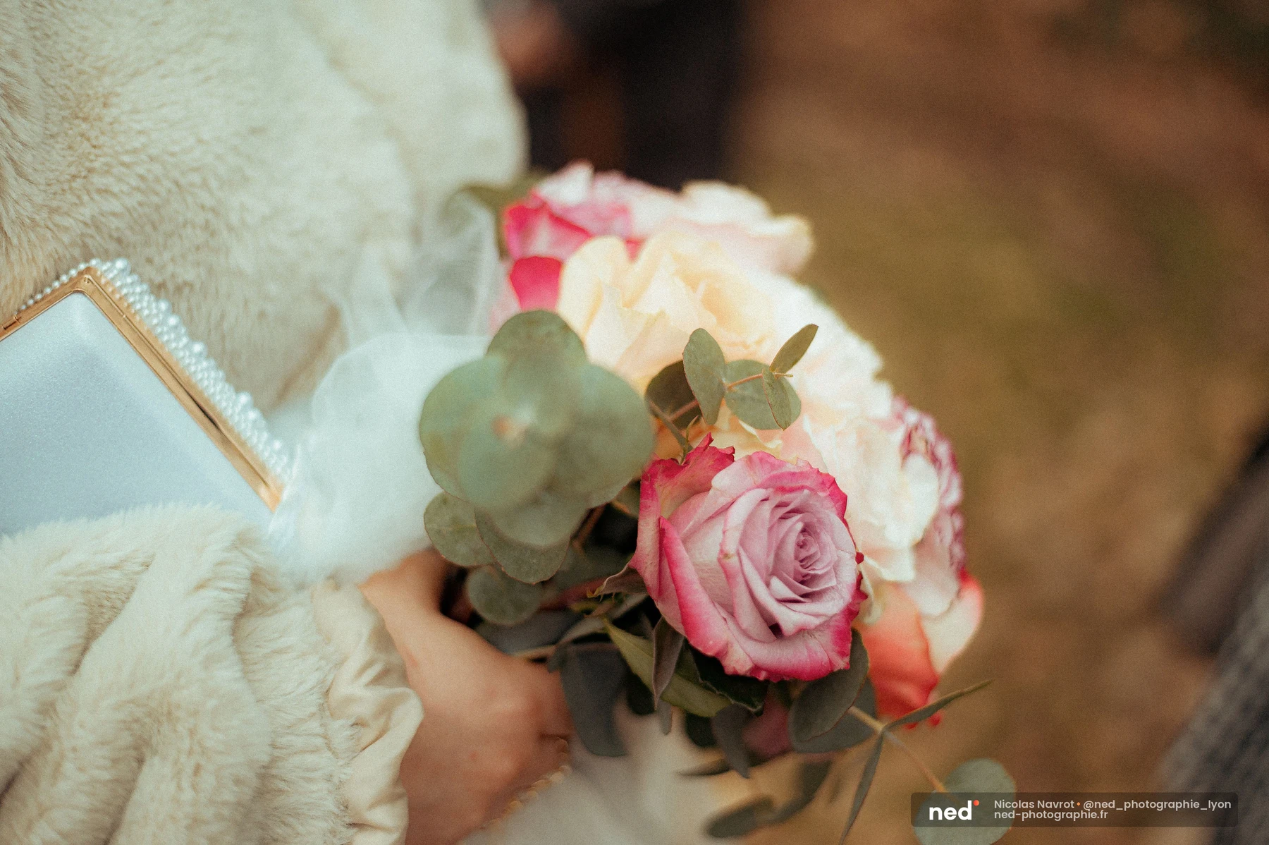 Bouquet de roses roses et pêche avec eucalyptus, tenu contre une veste en fourrure crème, à côté d'un clutch doré à détails de perles.