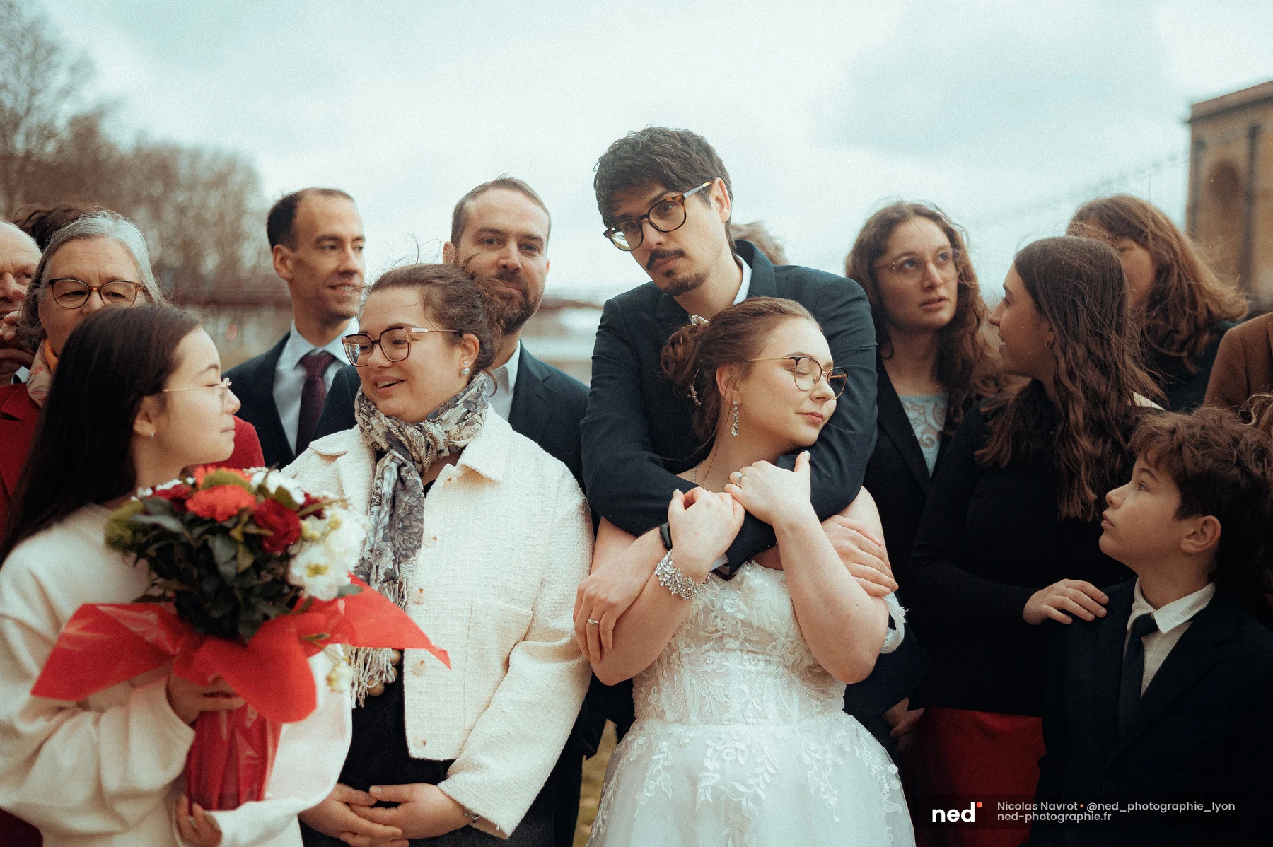 Le marié enlace la mariée par derrière au milieu du groupe de proches serrés, bouquet de roses rouges visible au premier plan.