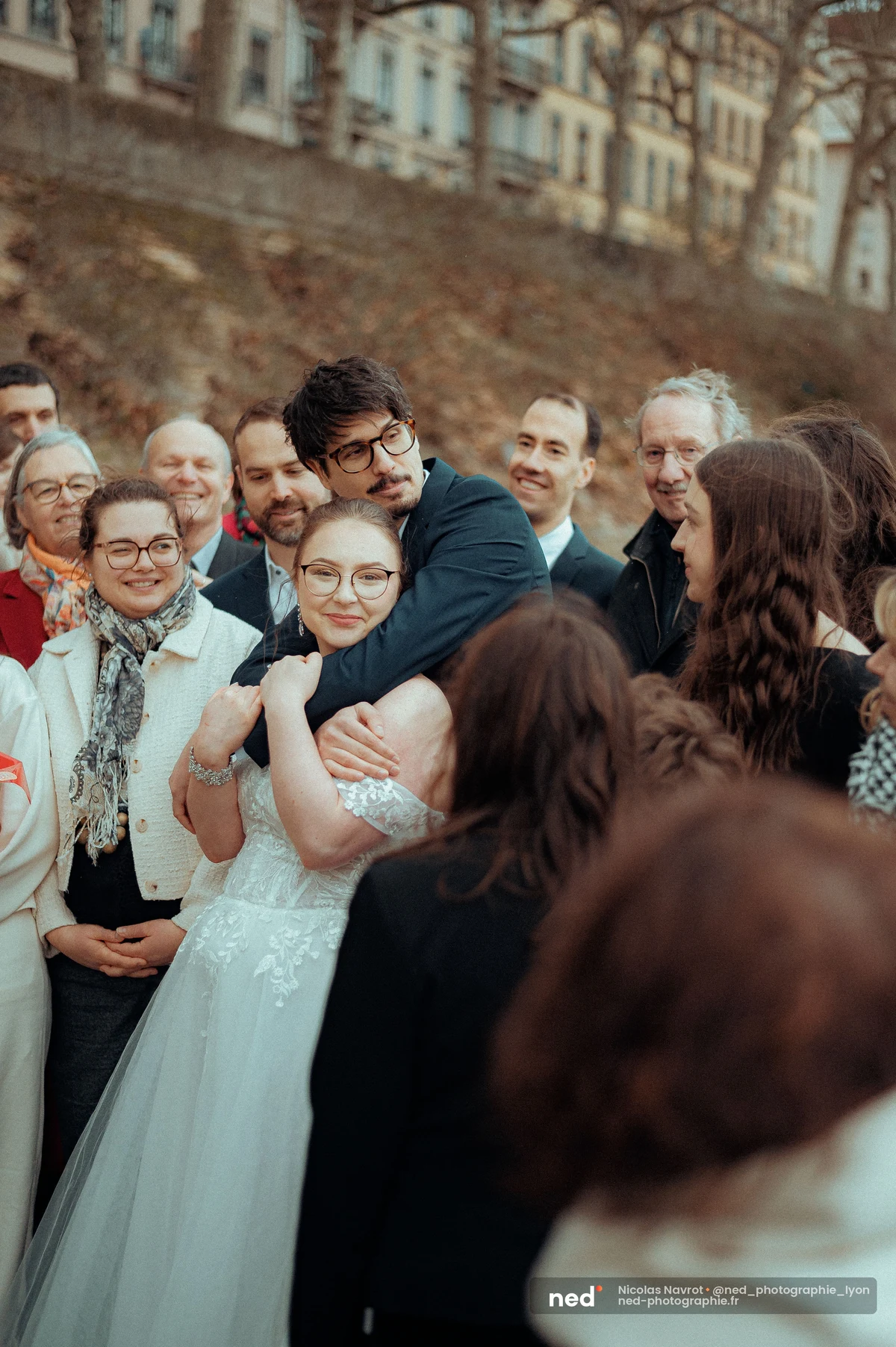 La mariée serre les bras du marié qui l'enlace, entourés de proches souriants, immeubles en pierre en arrière-plan.
