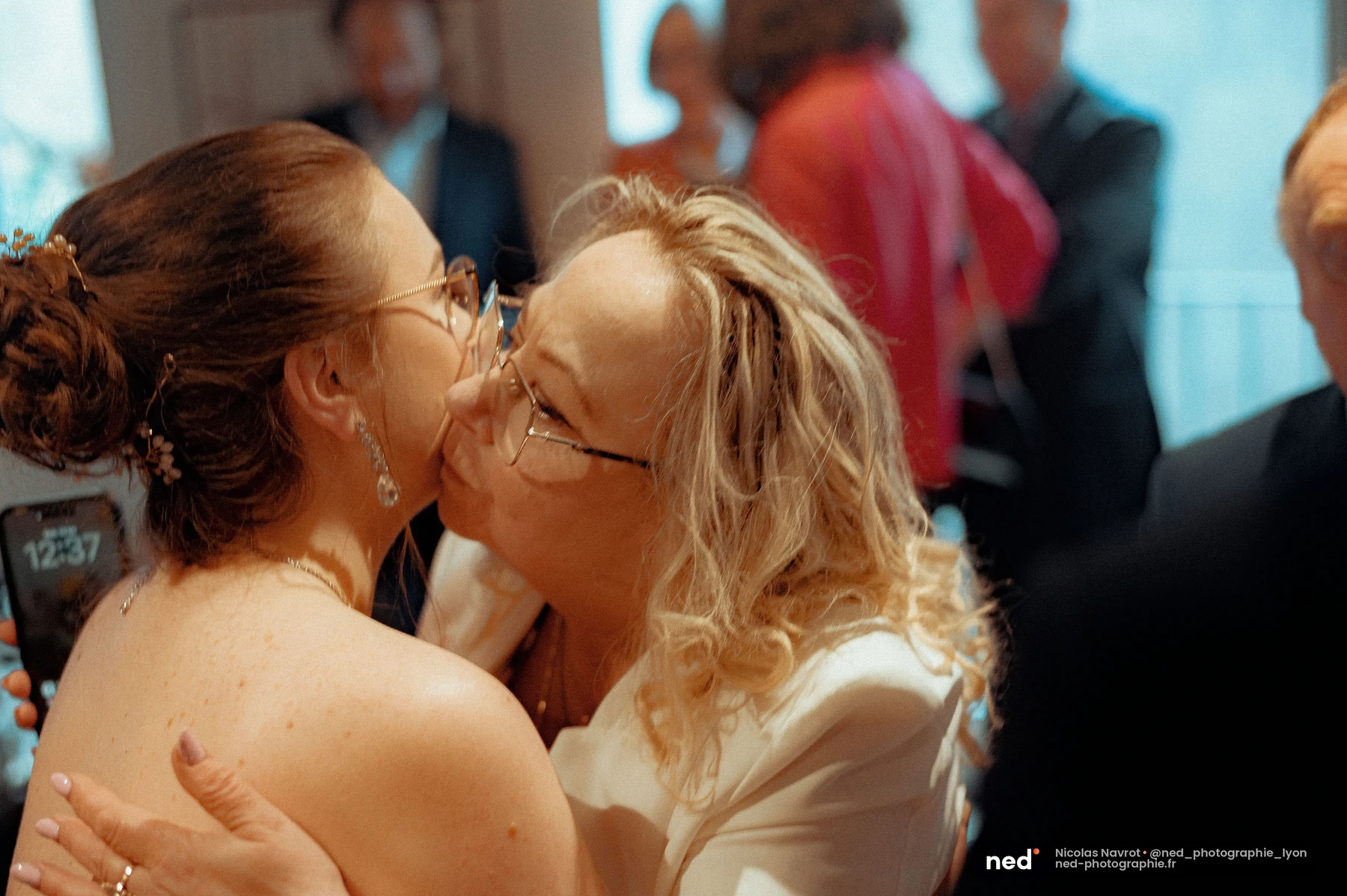 Une femme aux cheveux blonds bouclés embrasse la mariée sur la joue lors du cocktail, entourée de convives en arrière-plan flou.