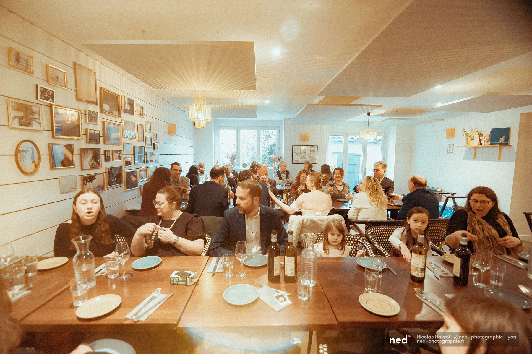 Salle de réception aux murs blancs couverts de cadres, convives assis à de longues tables en bois garnies de vaisselle et de bouteilles de vin.