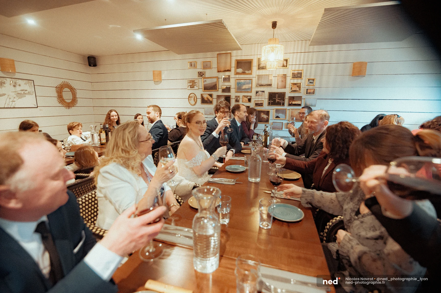 Convives levant leurs verres à vin lors d'un toast, mariée en robe blanche assise au centre de la tablée, salle décorée de cadres dorés.