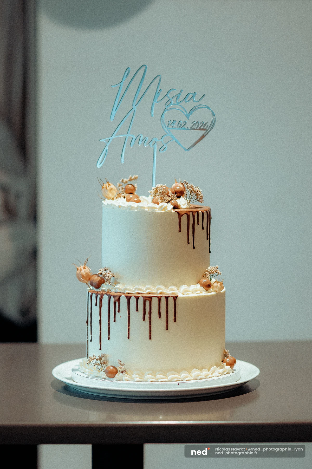 Gâteau de mariage à deux étages, glaçage blanc avec coulures de chocolat, décoré de fleurs séchées et d'un topper acrylique bleu gravé.