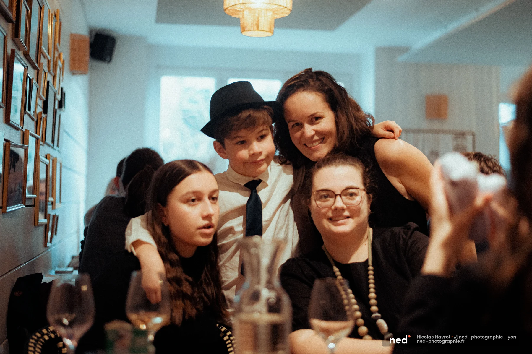 Groupe de quatre convives souriant autour d'une table de réception, dont un enfant en chemise blanche et cravate coiffé d'un chapeau noir.