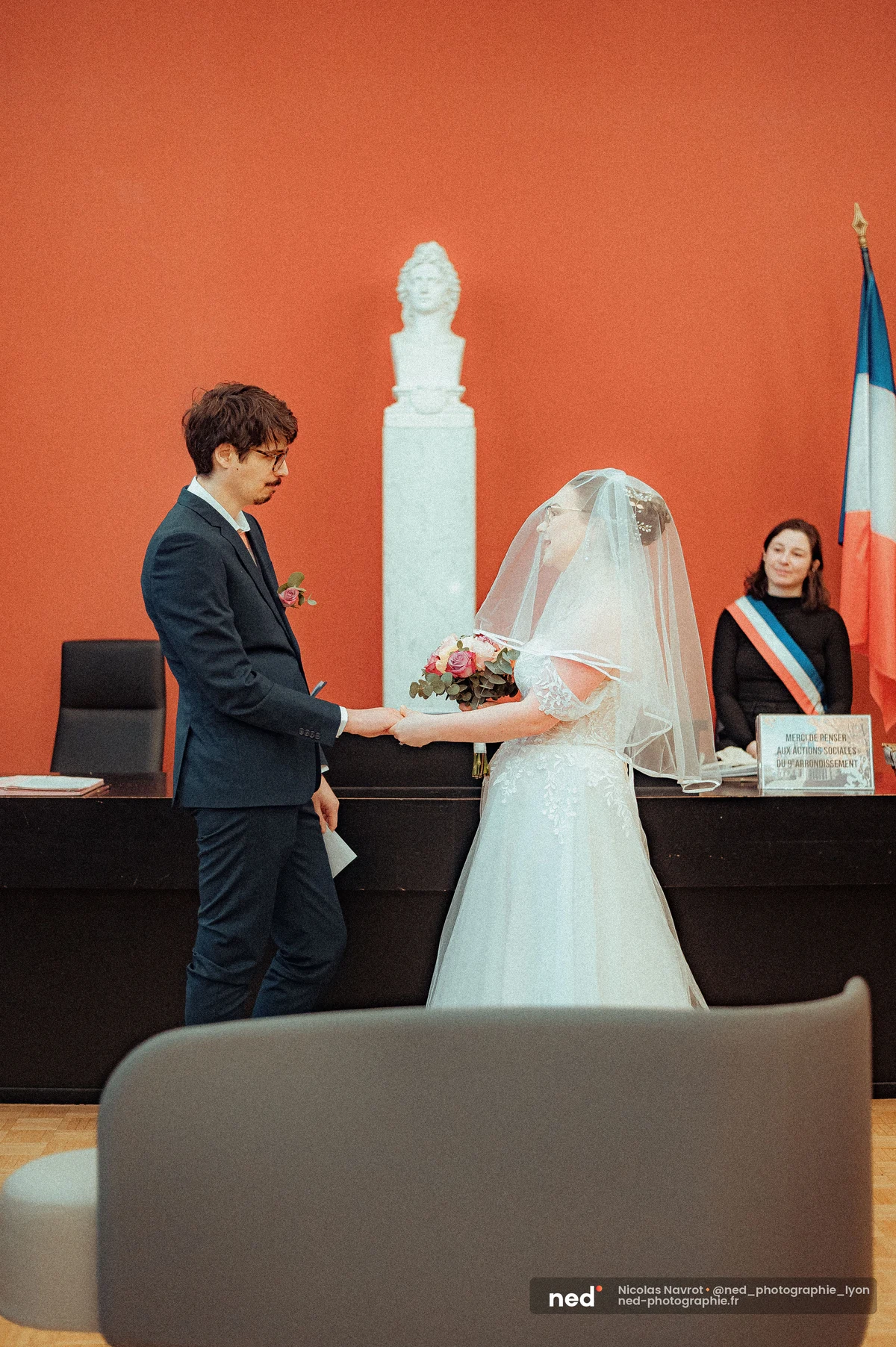 Mariés se tenant la main face à face devant l'officière d'état civil, salle aux murs rouge brique avec buste sculpté et drapeau tricolore.