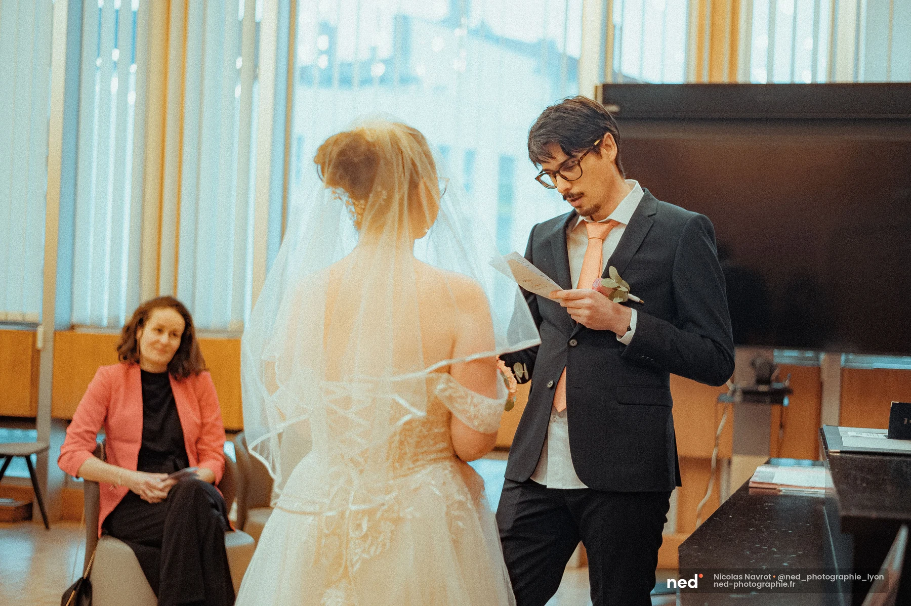 Le marié lit ses vœux depuis un carton manuscrit, face à la mariée de dos voilée, une invitée en veste corail visible en arrière-plan.