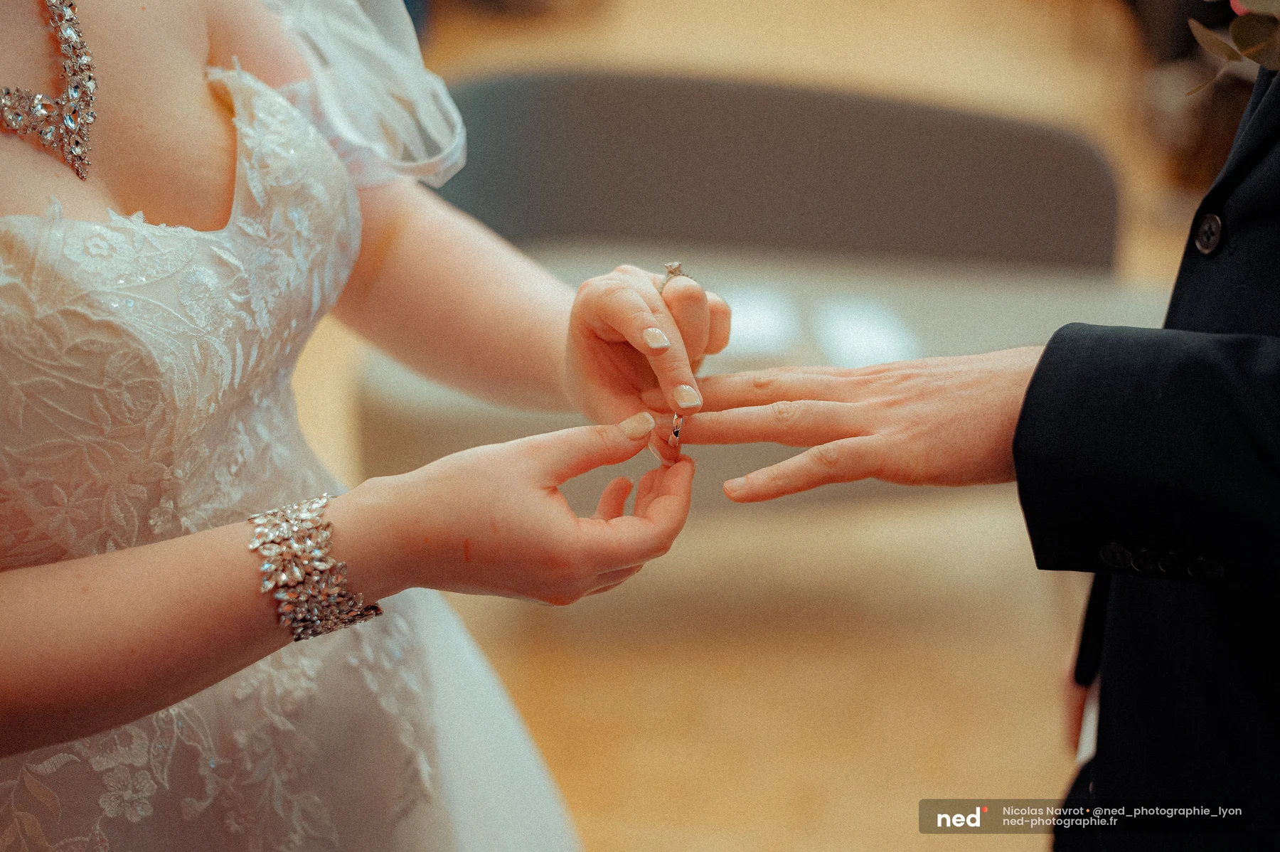 La mariée, bracelet strass au poignet, passe un anneau au doigt du marié ; robe brodée visible à gauche, fond flou de la salle.