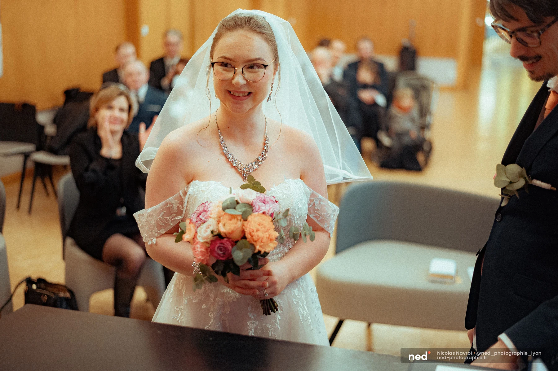 Mariée en robe dentelle, voile blanc et collier strass, tenant un bouquet de roses orangées et roses, souriante face à l'objectif dans une salle de mairie.
