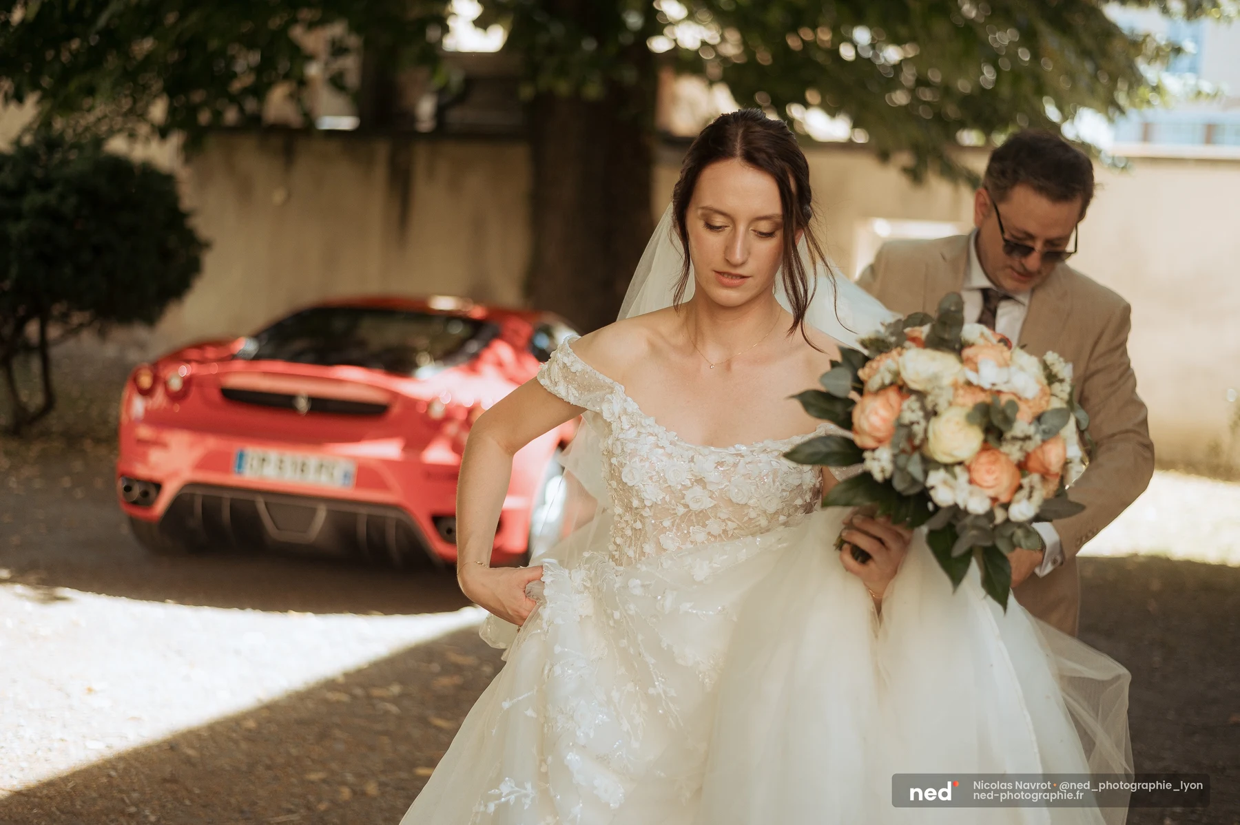 Mariée en robe princesse off-shoulder avec bouquet pêche et blanc, aidée par un proche pour porter sa jupe, devant une voiture de sport rouge à l'extérieur.