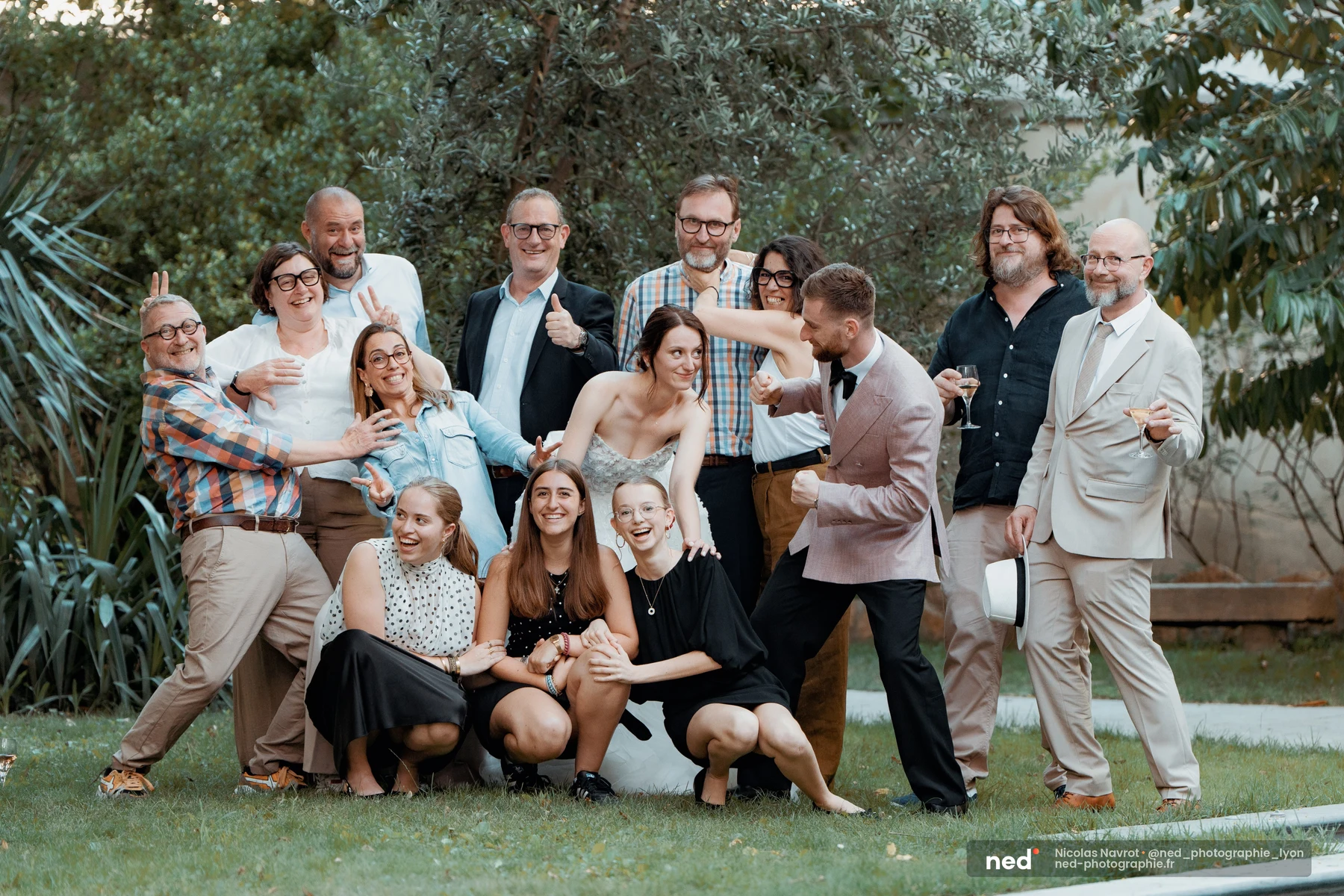 Groupe d'une quinzaine d'invités posant en plein air avec la mariée en robe blanche au centre ; certains font des gestes espiègles, tous sourient.