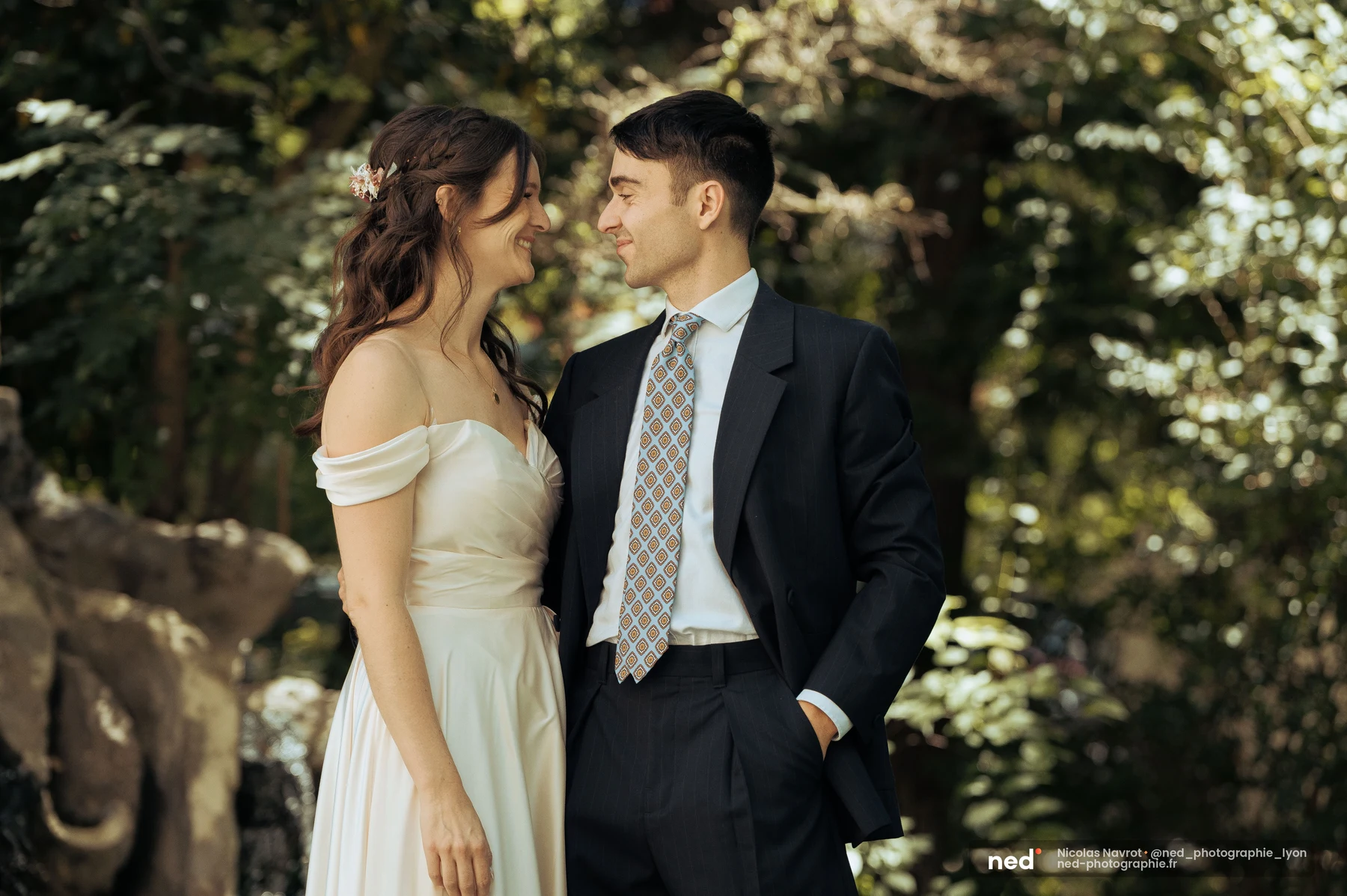 Couple d'invités se regardant de près dans un jardin verdoyant et ensoleillé : robe blanche off-shoulder pour elle, costume sombre et cravate à motifs pour lui.