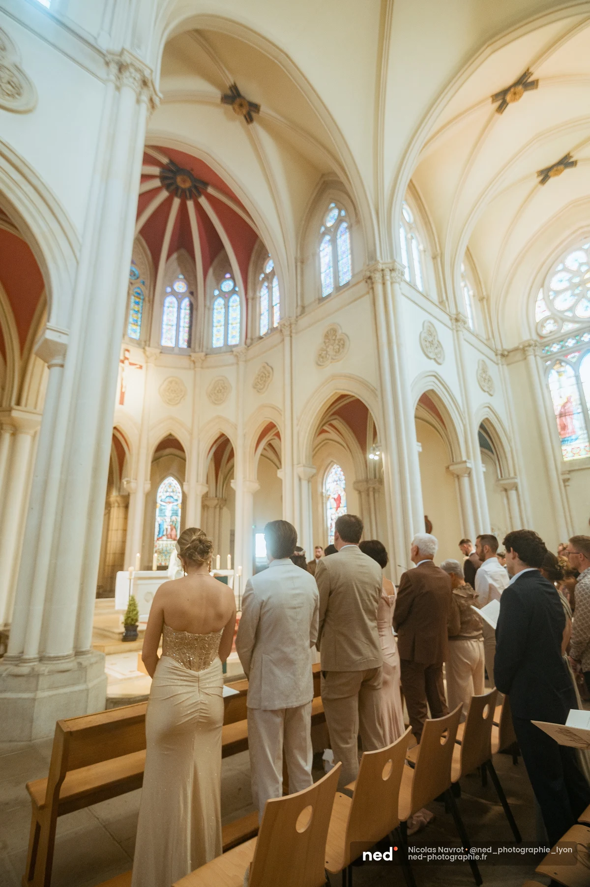 Vue de la nef d'une église gothique pendant la cérémonie de mariage : invités debout face à l'autel, voûtes blanches, vitraux colorés et coupole rouge.