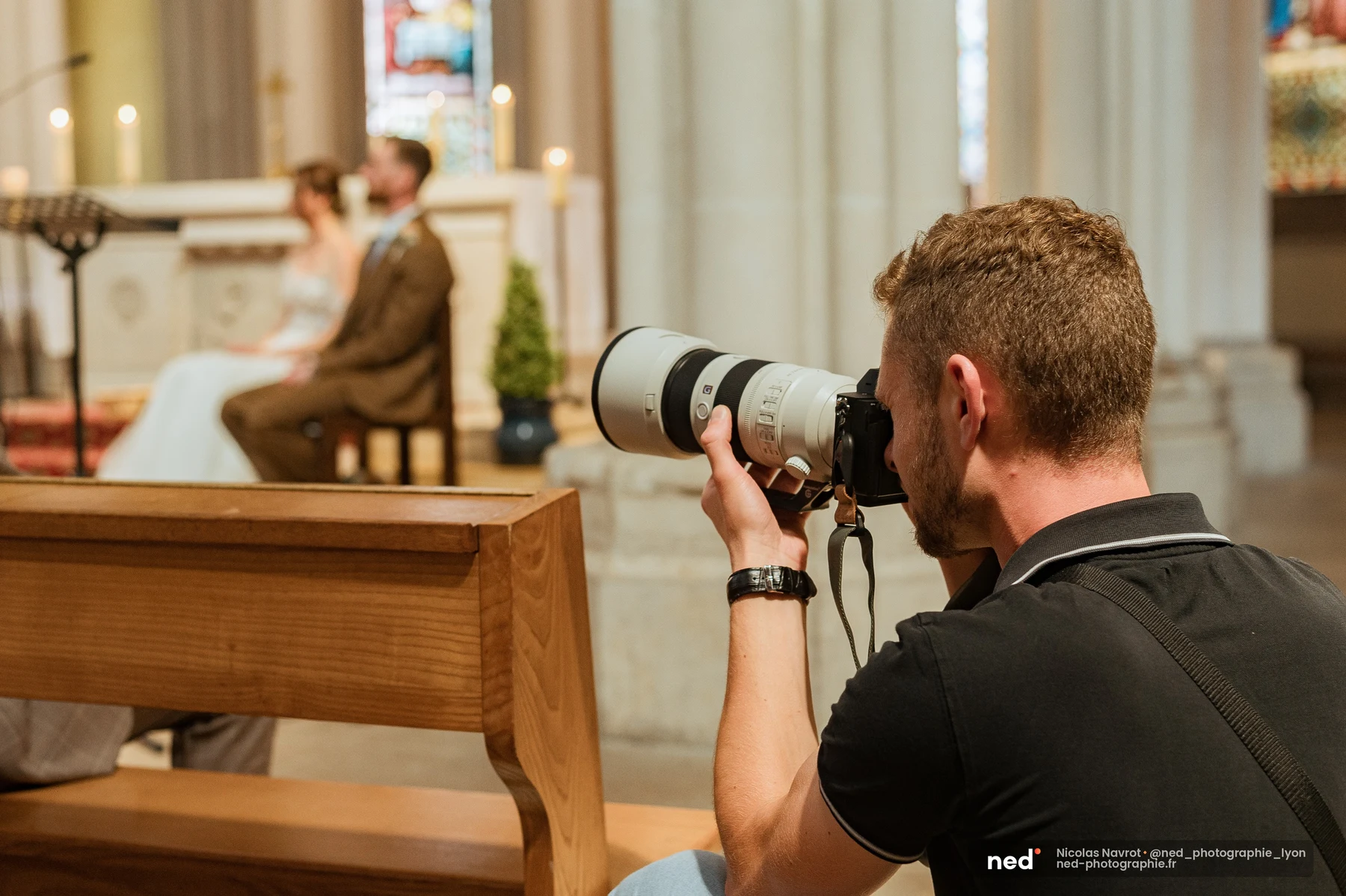 Photographe de mariage accroupi dans la nef, téléobjectif blanc pointé vers les mariés assis à l'autel en arrière-plan flou, pendant la cérémonie religieuse.