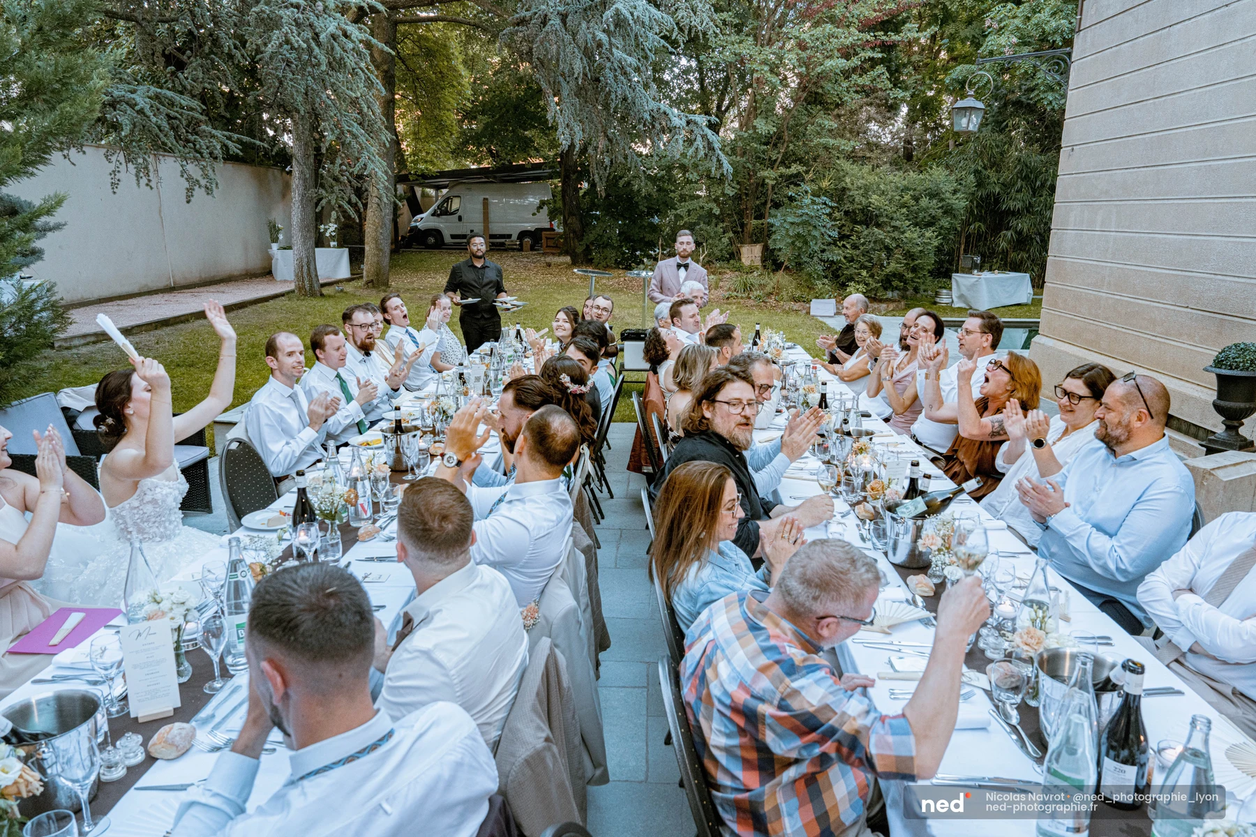 Vue plongeante sur une longue table de mariage en extérieur : une trentaine d'invités applaudissent en direction d'un orateur debout au fond du jardin.