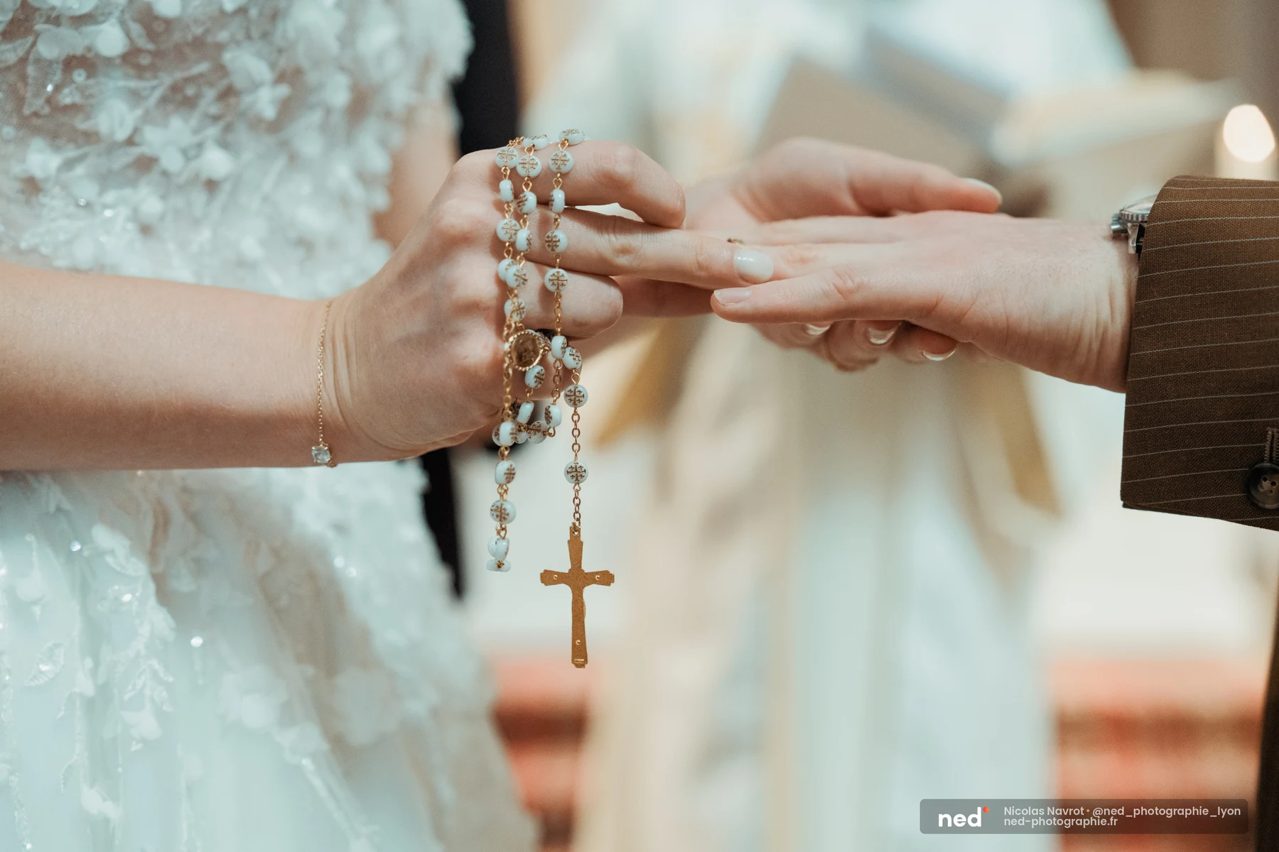 Gros plan sur deux mains échangeant une alliance lors d'une cérémonie religieuse ; un chapelet doré aux perles bleues entoure les doigts de la mariée.