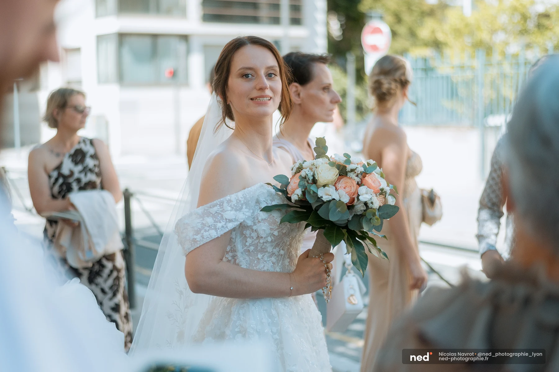 Mariée en robe dentelle off-shoulder tenant son bouquet pêche et blanc, regardant vers l'objectif entourée d'invités flous au soleil.
