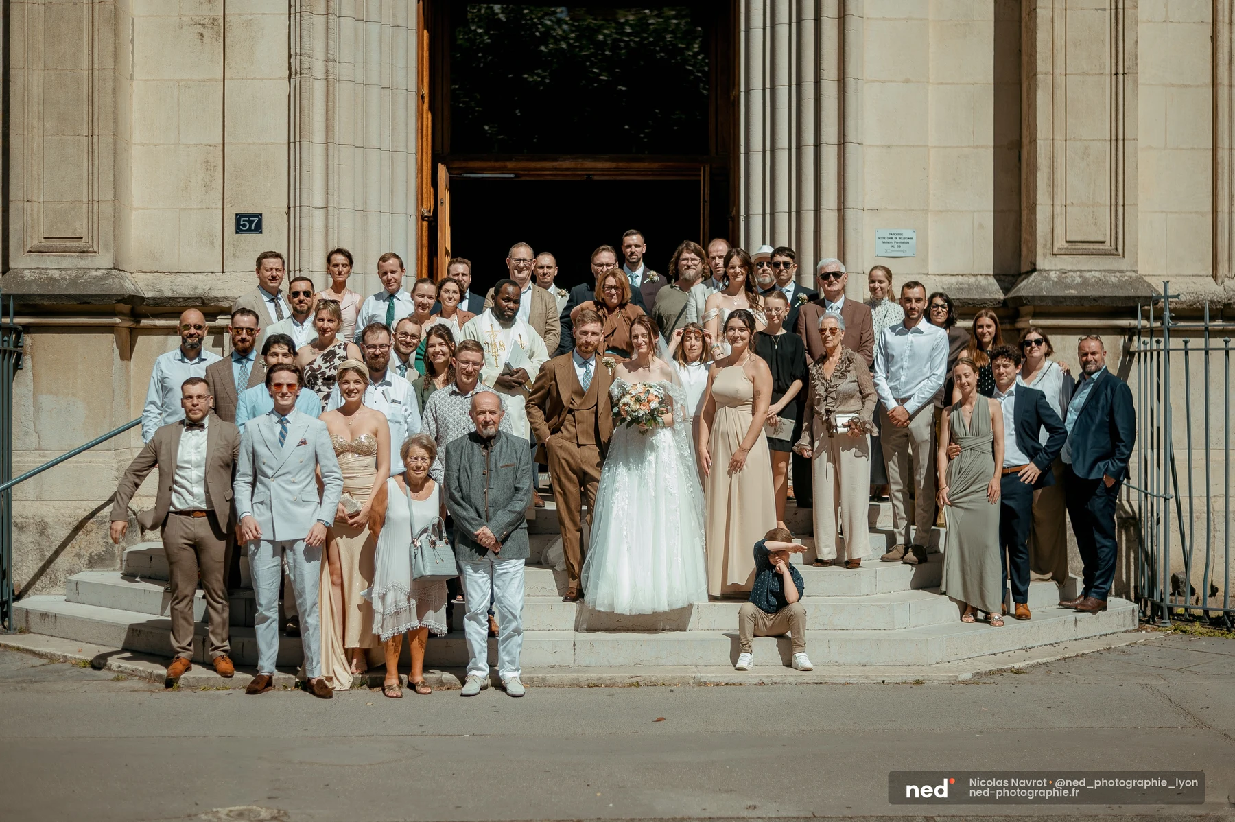 Photo de groupe sur les marches d'une église à colonnes ; mariés en robe blanche et costume marron au centre, entourés d'une trentaine d'invités.
