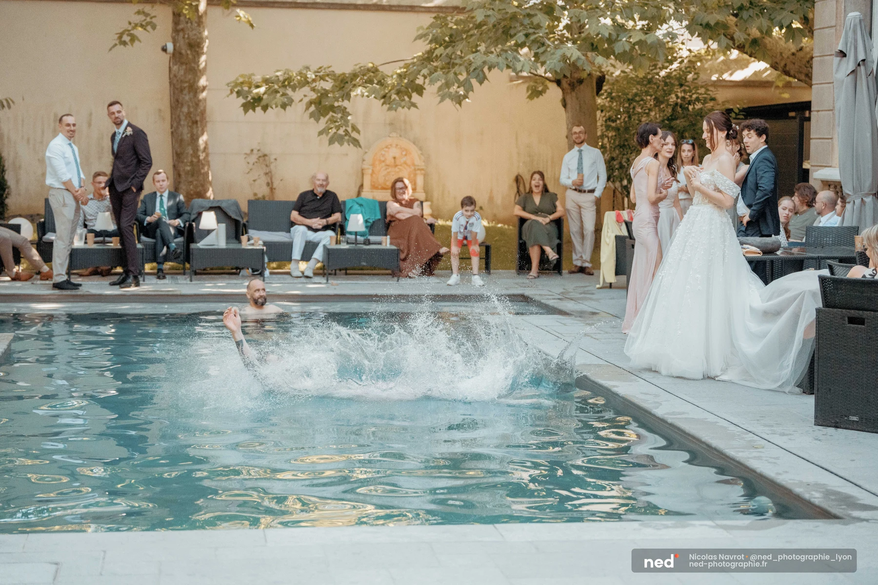 Invité ayant plongé habillé dans une piscine lors d'une réception de mariage en extérieur ; la mariée en robe blanche regarde la scène depuis le bord.