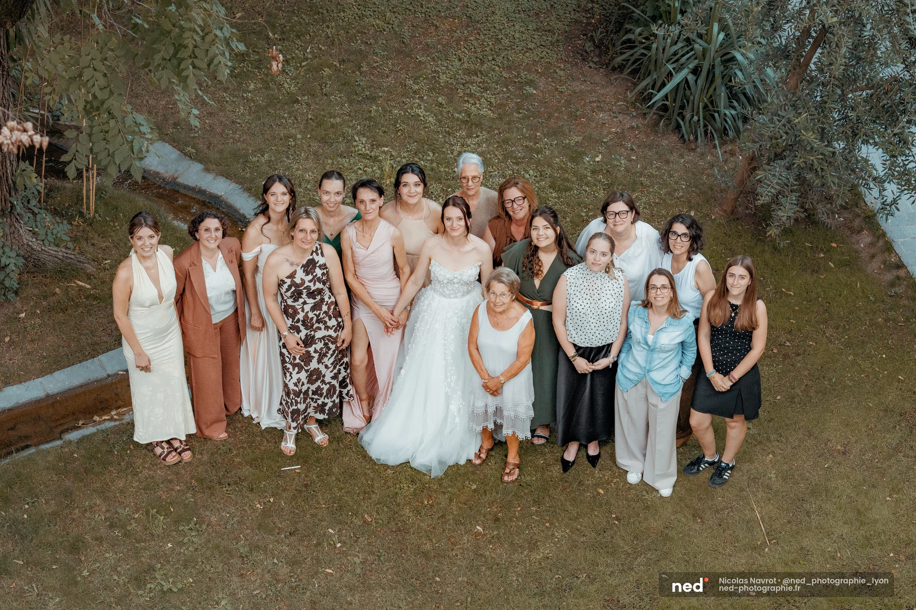 Photo aérienne de la mariée en robe blanche entourée d'une quinzaine d'invitées sur une pelouse, rangées sur deux niveaux face à l'objectif.