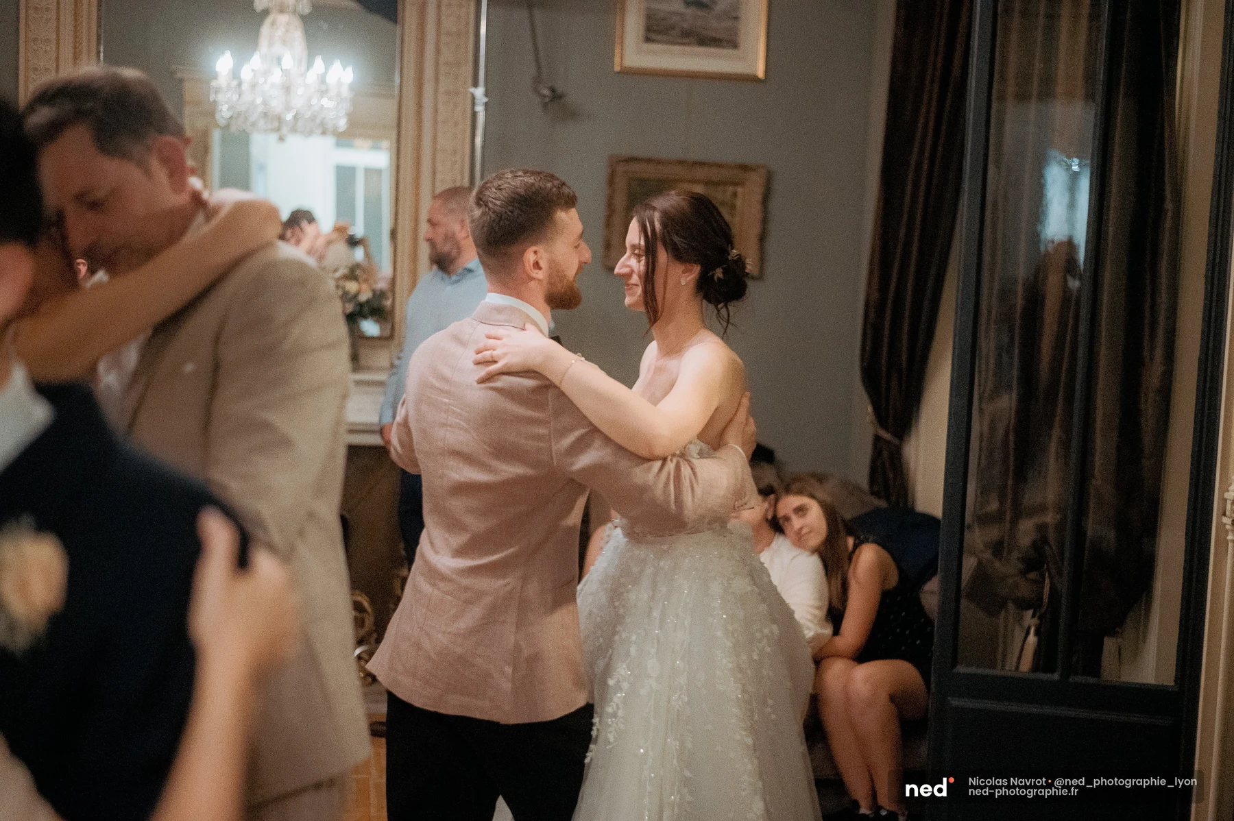 Mariée en robe blanche tulle et marié en veste rose dansent enlacés dans un salon bourgeois ; un grand miroir doré reflète la salle derrière eux.