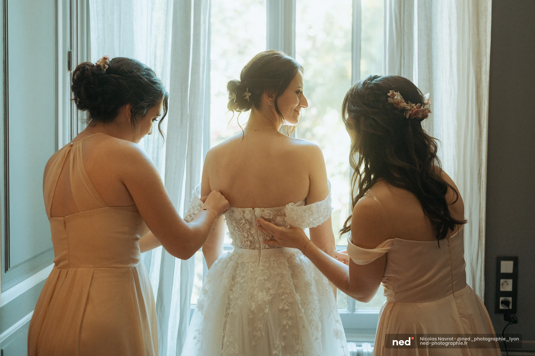 Deux demoiselles en rose poudré boutonnent le dos de la robe en tulle et dentelle de la mariée, devant une fenêtre aux voilages lumineux.