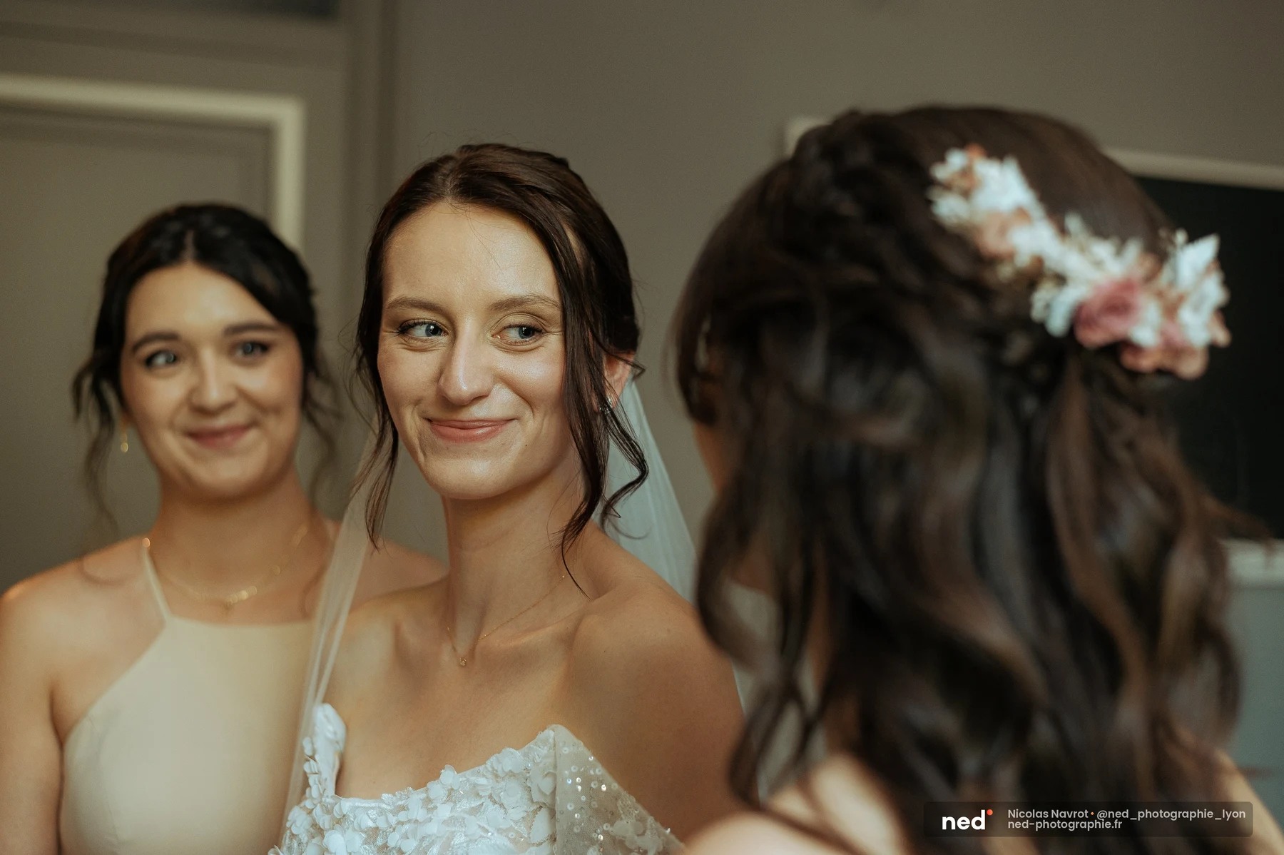 Mariée en robe off-shoulder à appliqués floraux et voile blanc, souriant à une proche pendant les préparatifs, demoiselle d'honneur de dos au premier plan.
