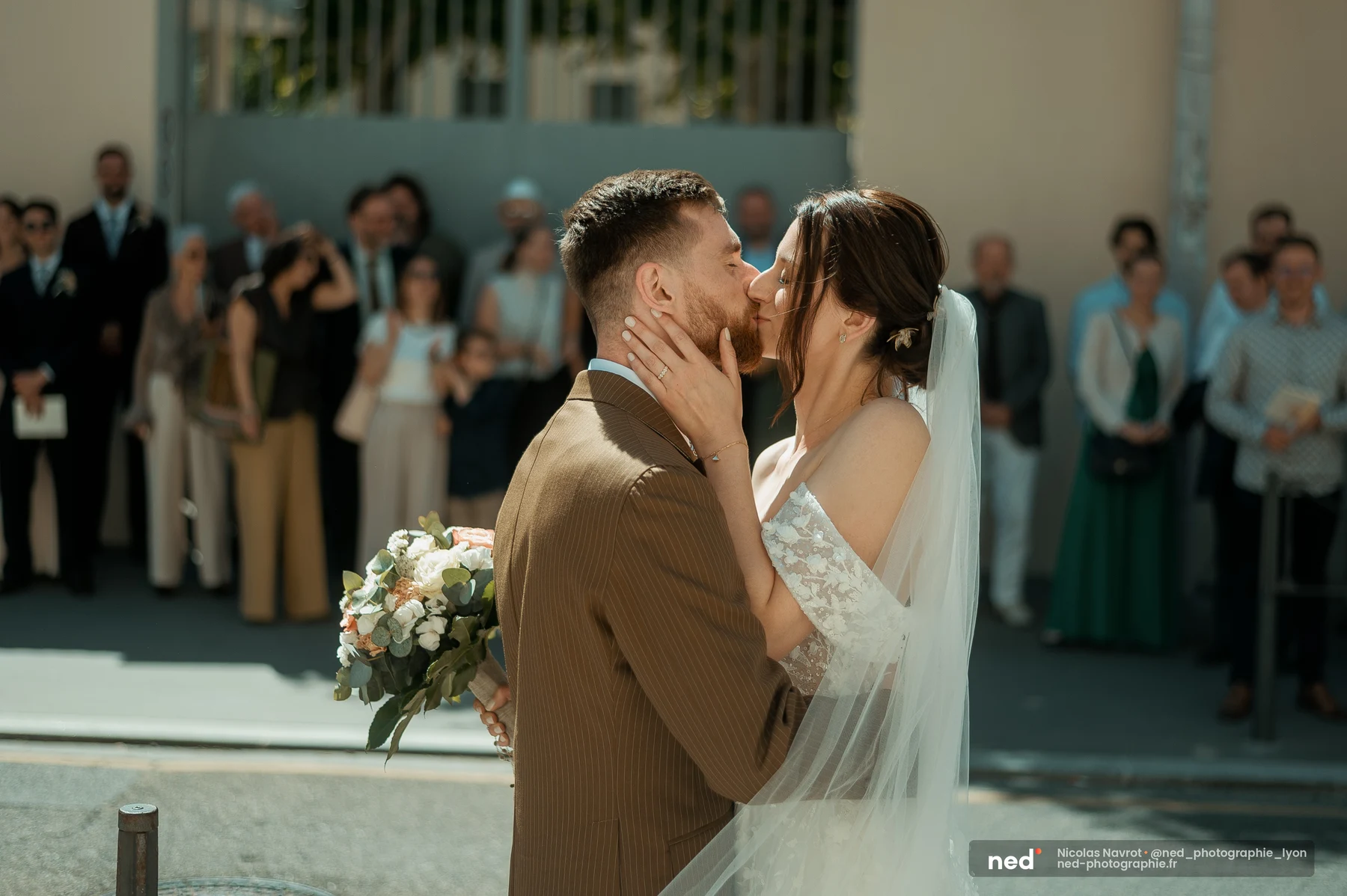 Couple de mariés s'embrassant sur un parvis ensoleillé, entouré de proches floutés ; la mariée tient la joue du marié, voile au vent.
