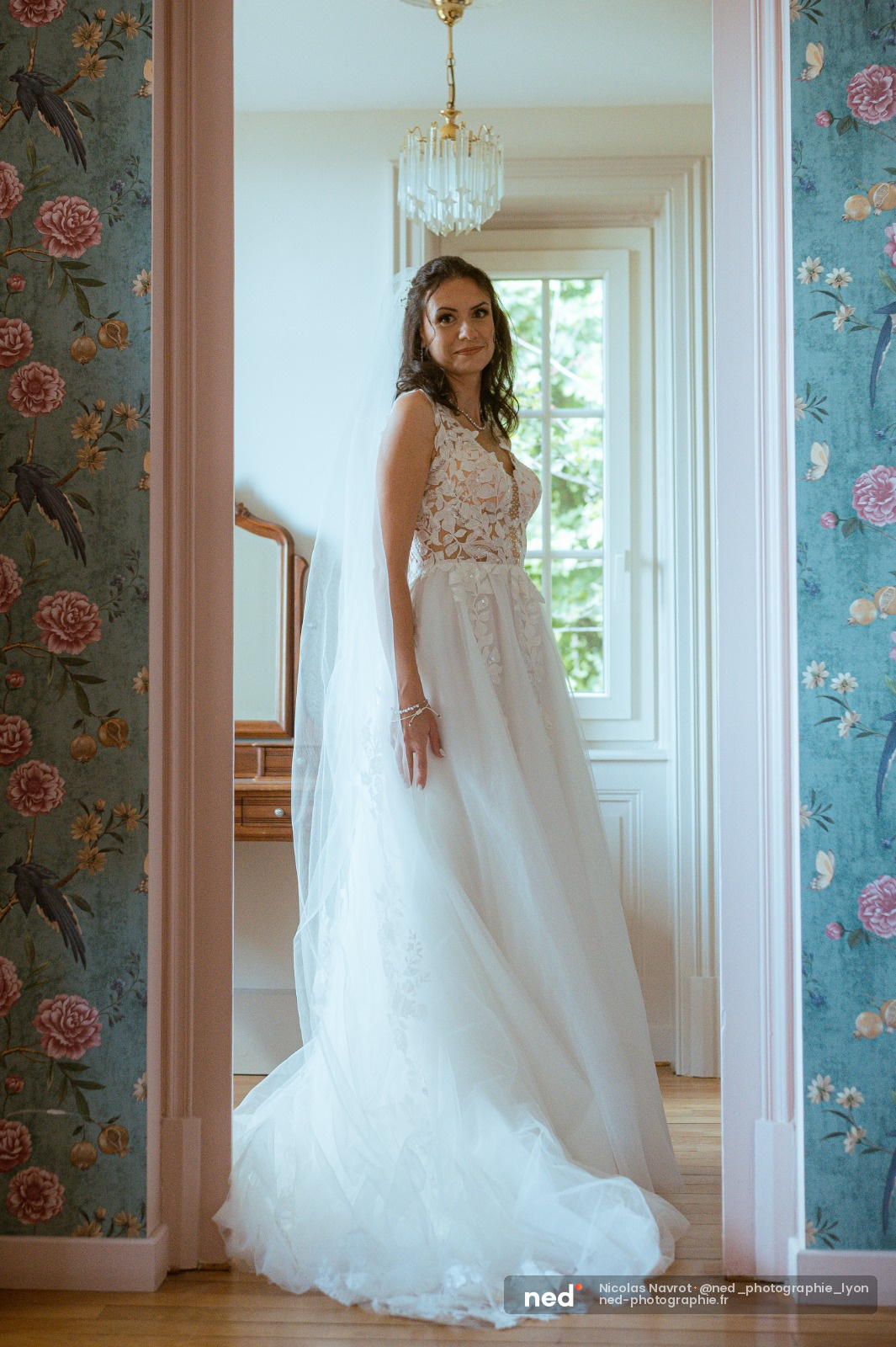 Mariée en robe de mariage blanche avec bustier en dentelle se tourne dans un couloir classique aux murs bleu-vert, lustre cristal au plafond
