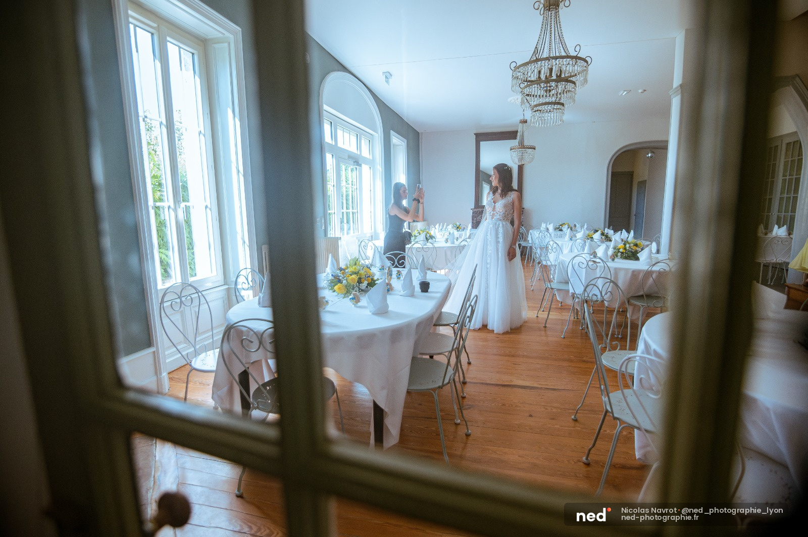 Mariée en robe blanche debout dans une salle de réception élégante avec tables rondes blanches, vue depuis une porte vitrée